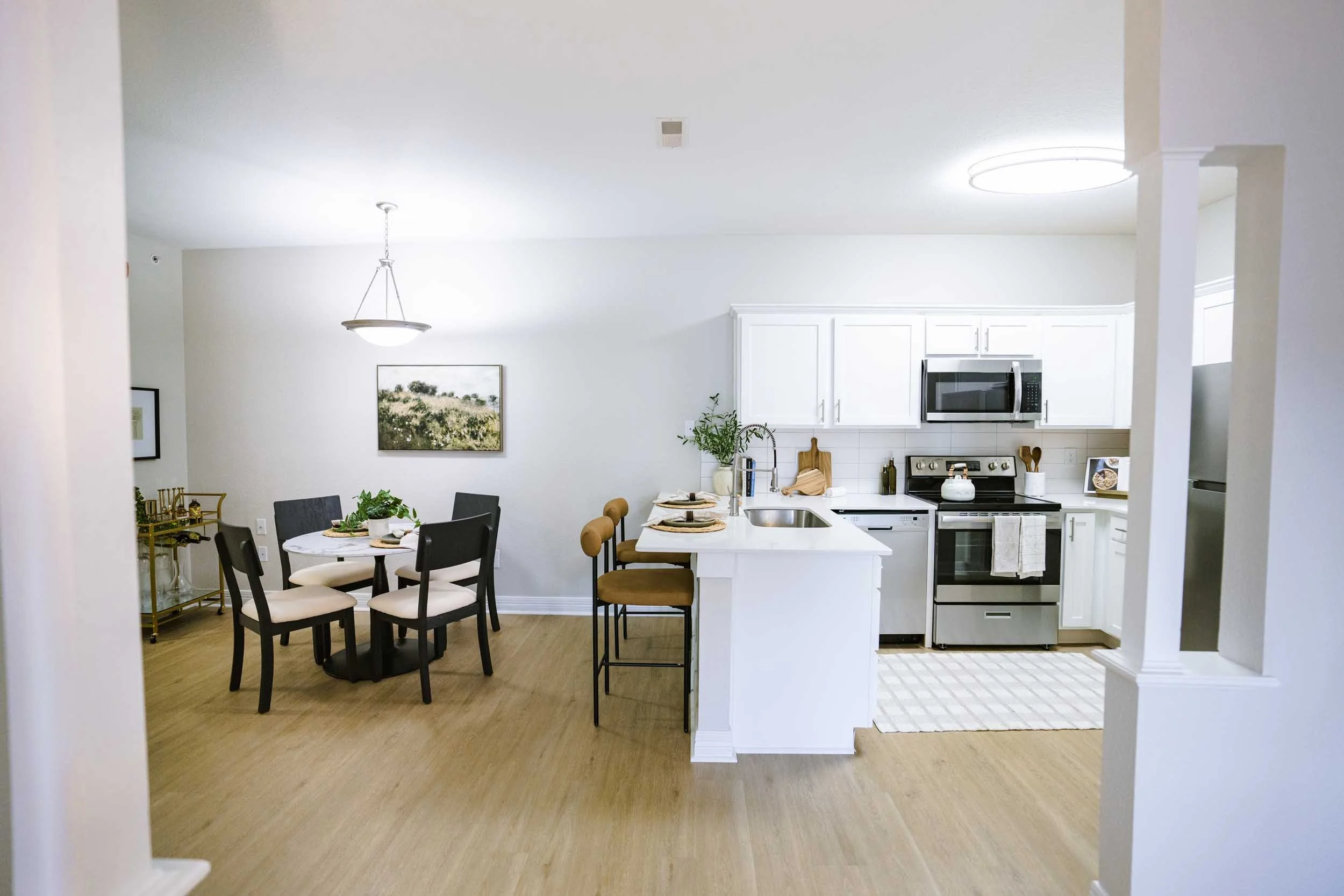 Open-concept kitchen and dining area with white cabinetry and black appliances, a round dining table with six chairs, and a wooden barstool at the kitchen island. Walls decorated with framed artwork and houseplants are visible in the background.