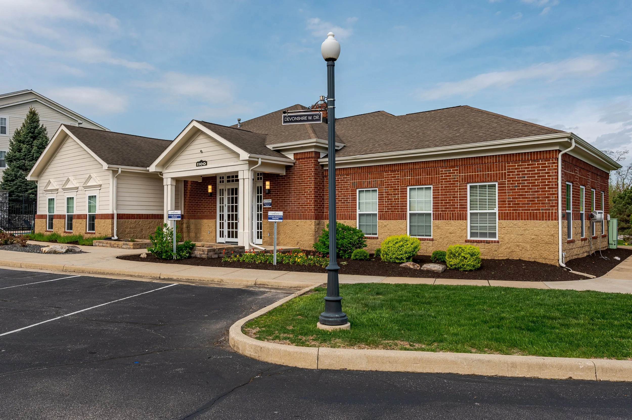 A single-story building with a mix of brick and beige siding, with white trim and a brown shingled roof, appears to be a professional or medical office. There is a parking lot in front with empty spaces, a lamppost with a street sign reading 'Devonsh