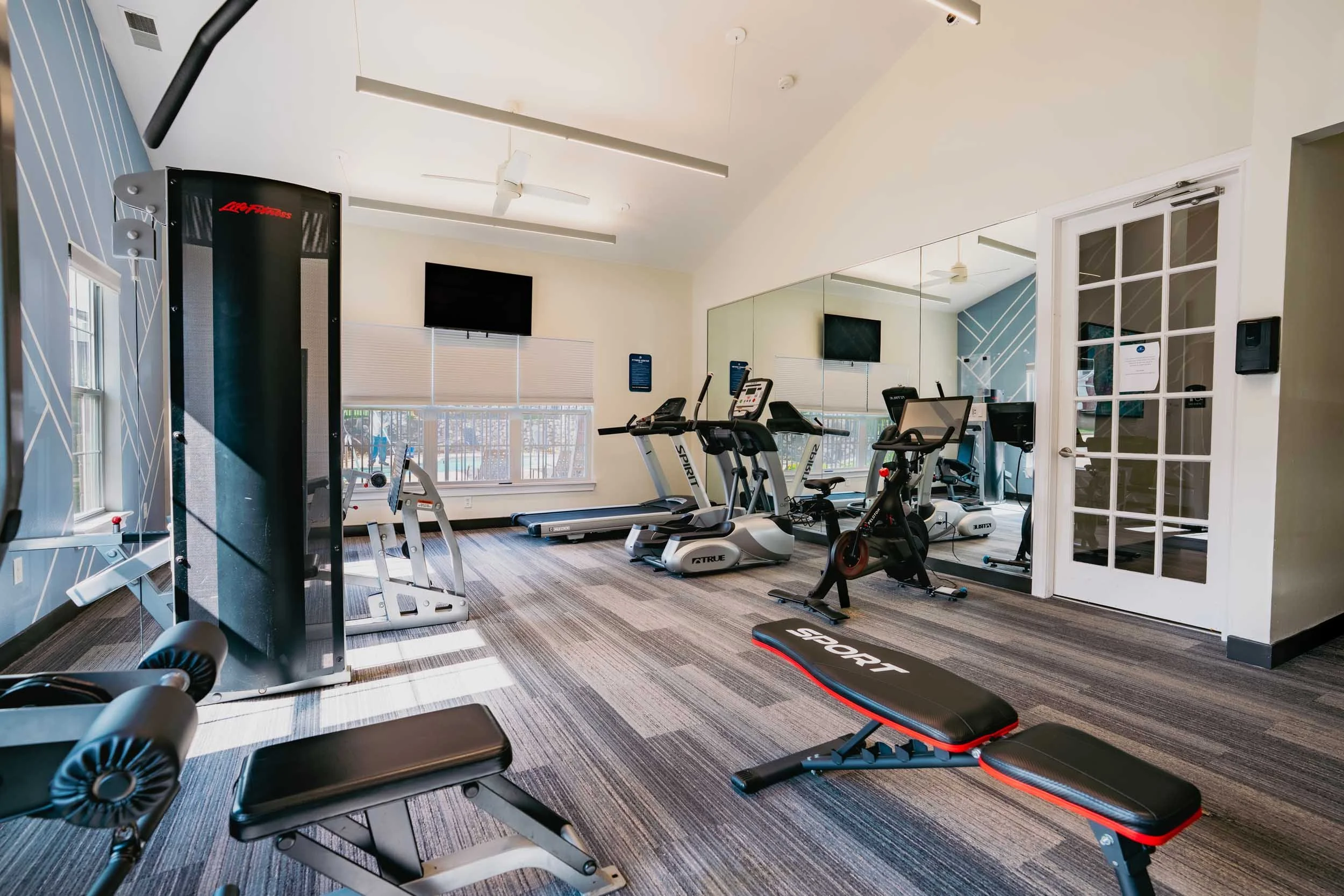 A gym room with exercise equipment including treadmills, exercise bikes, and a weight bench. The room has large windows with blinds, a wall-mounted TV, and mirrored walls.