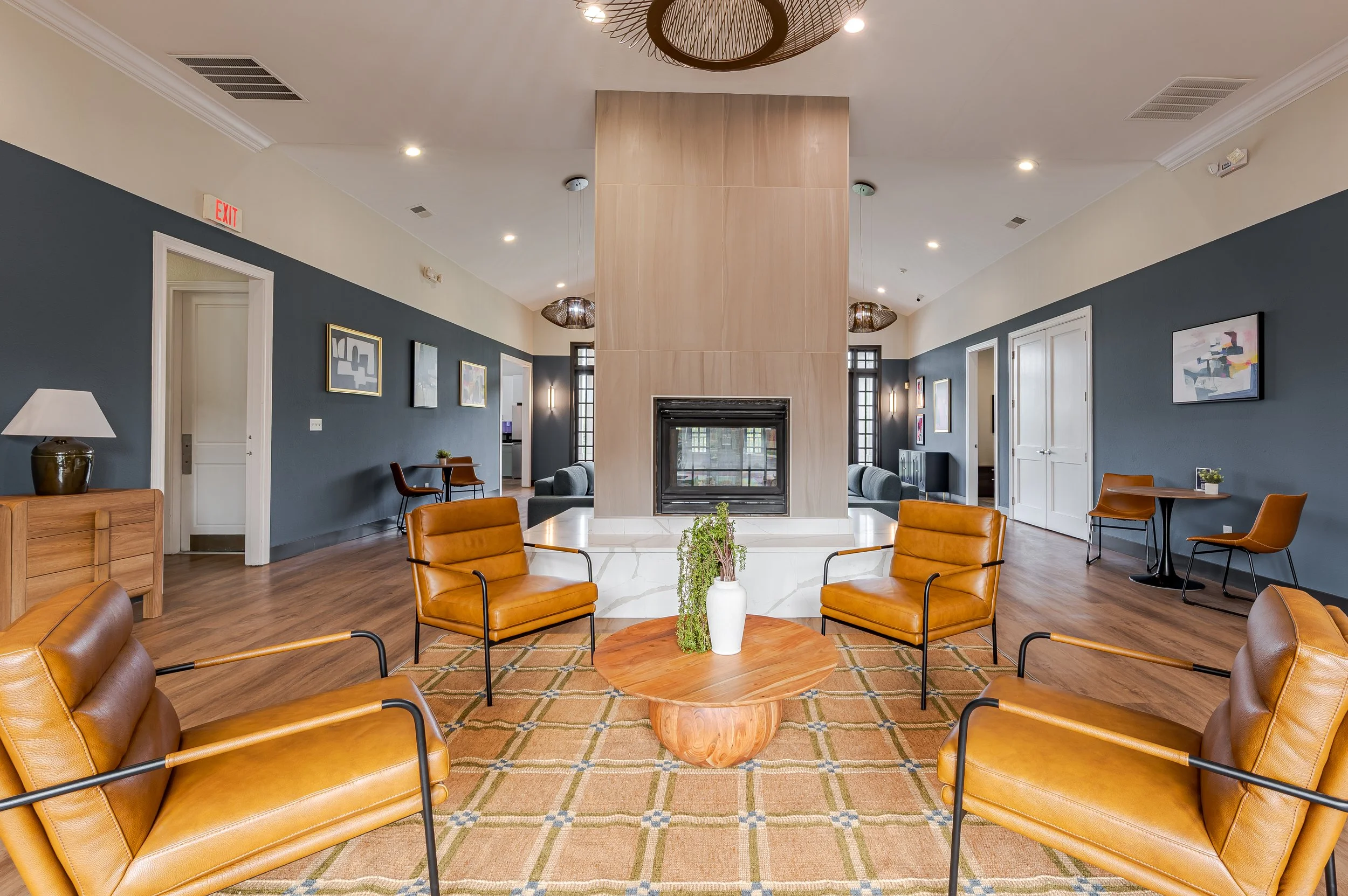 A modern living room with a central fireplace, surrounded by blue walls decorated with abstract art. Leather chairs and wooden tables are arranged around a white marble hearth, with a vase of plants on a wooden coffee table. The ceiling features rece