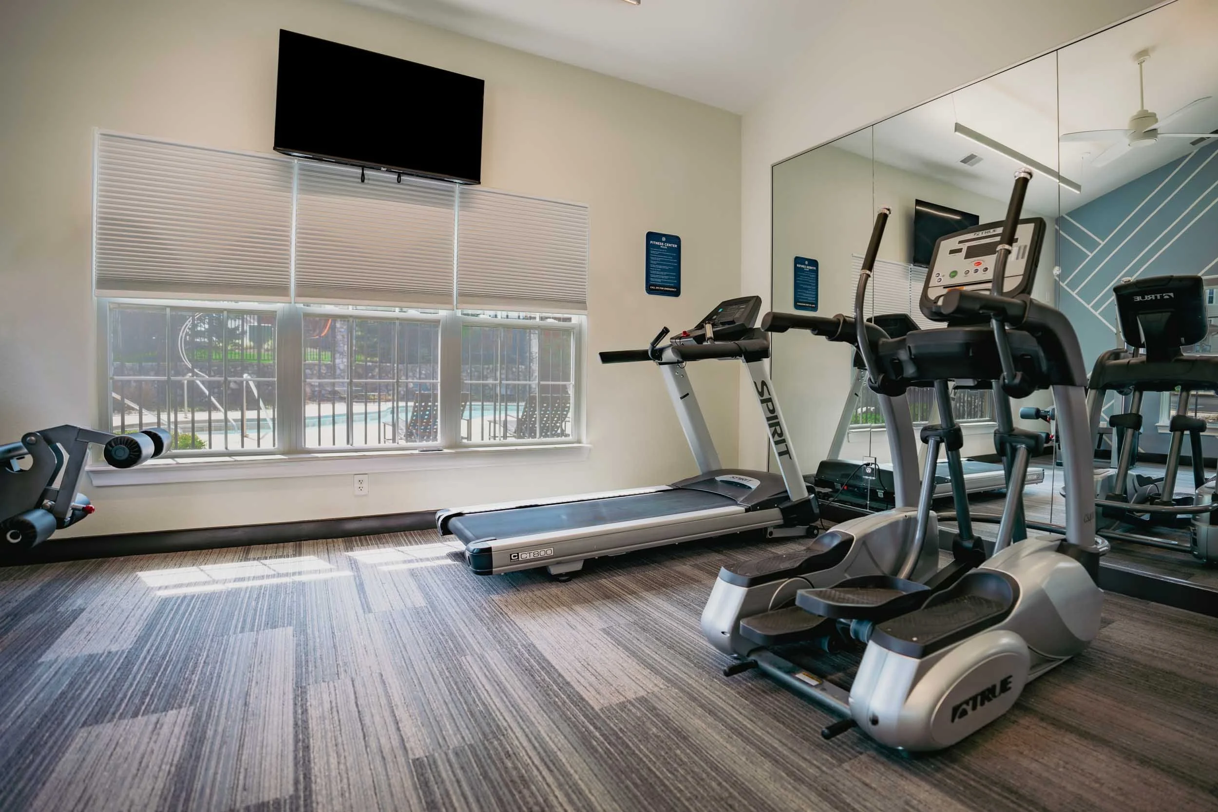 A gym room with a treadmill, elliptical machine, and a mirrored wall, with a large window showing an outdoor pool and patio area.