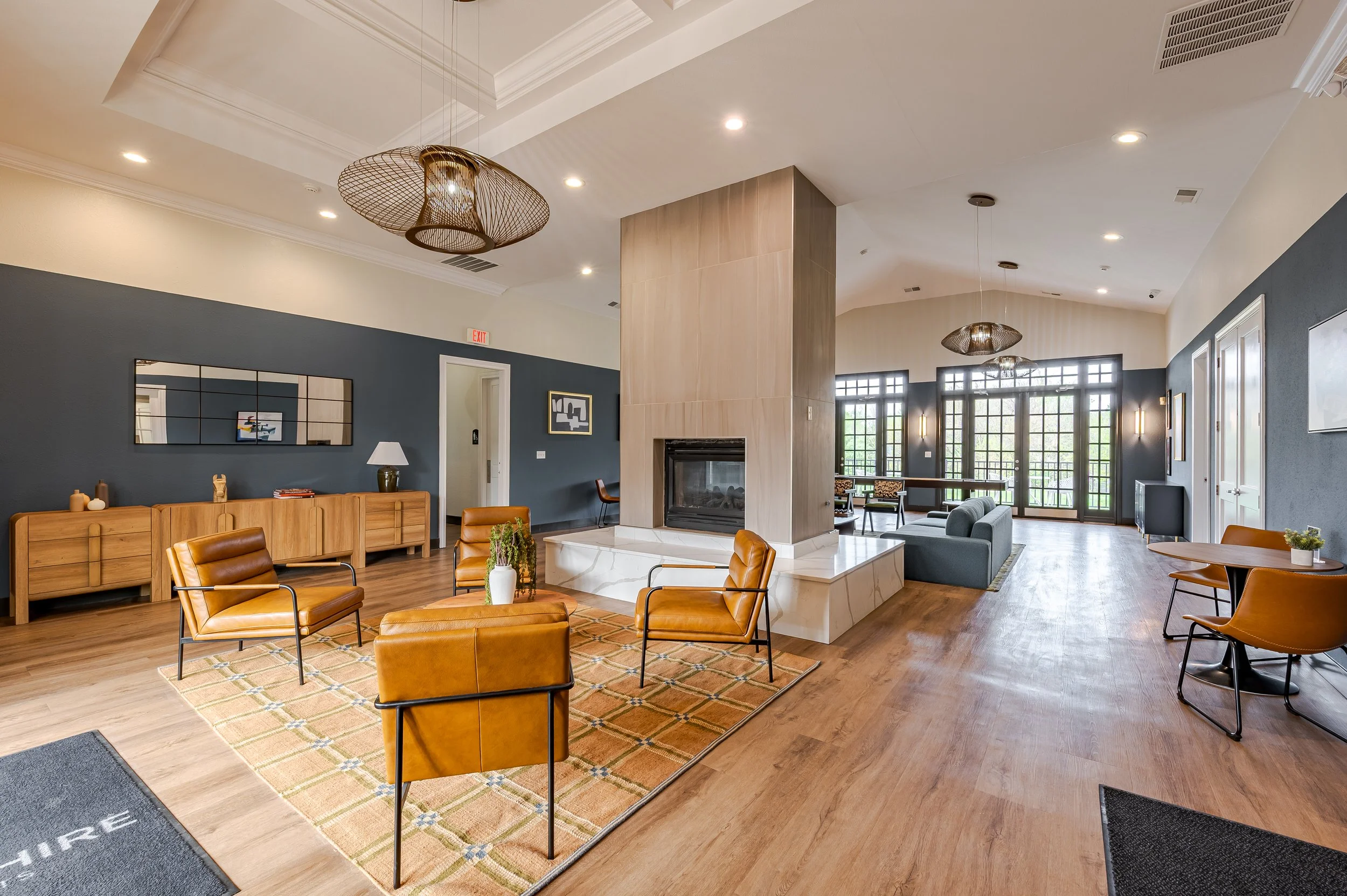A spacious, modern living room with a high ceiling, wooden flooring, and large windows. The room features a centered fireplace with a marble base, surrounded by leather and fabric chairs and a wooden sideboard. Hanging light fixtures and artwork are 