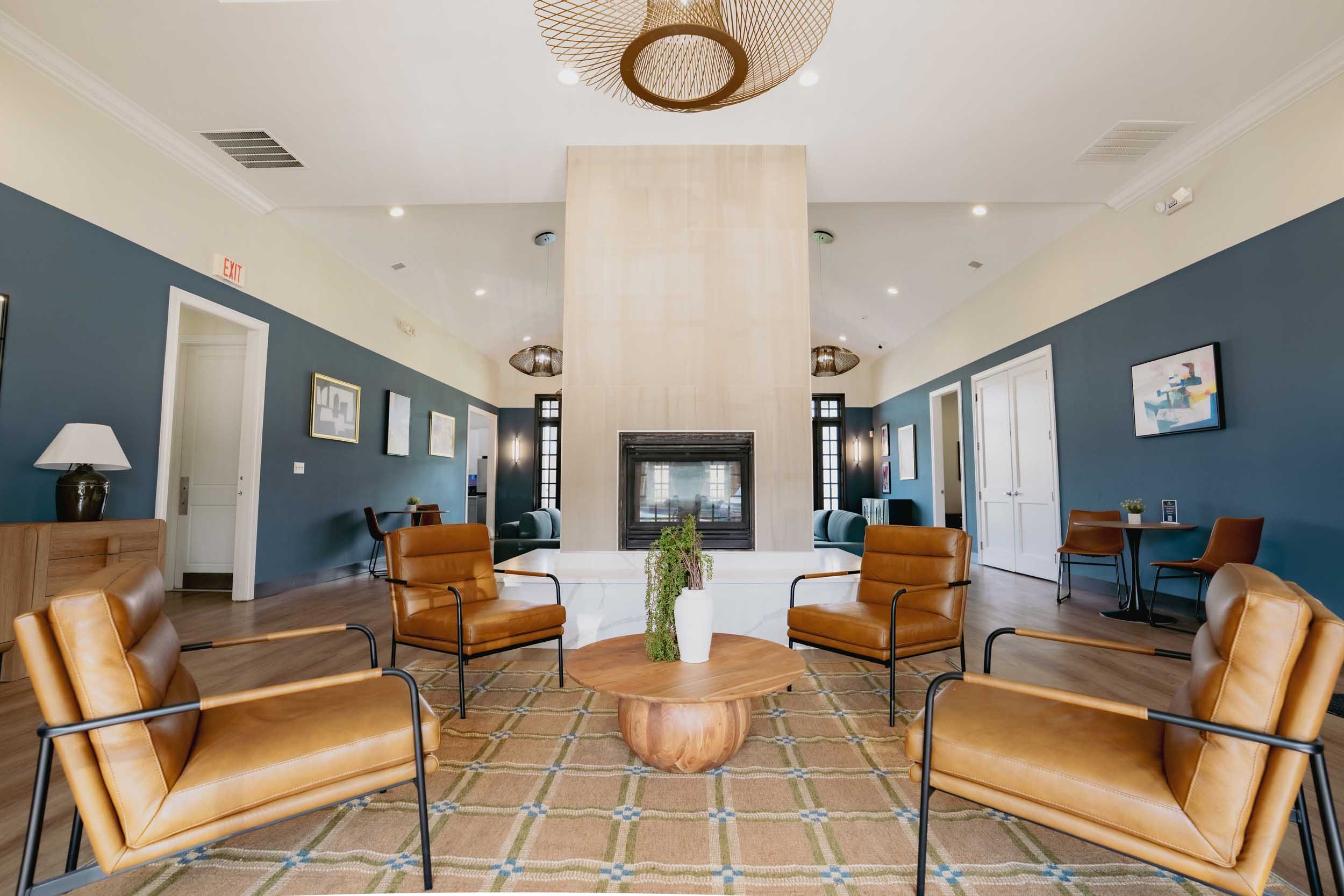 A spacious lounge area with a fireplace at the center, surrounded by tan leather chairs arranged in a circular pattern on a beige and green plaid rug. The room has blue walls adorned with framed artwork, and a table with a white vase holding greenery