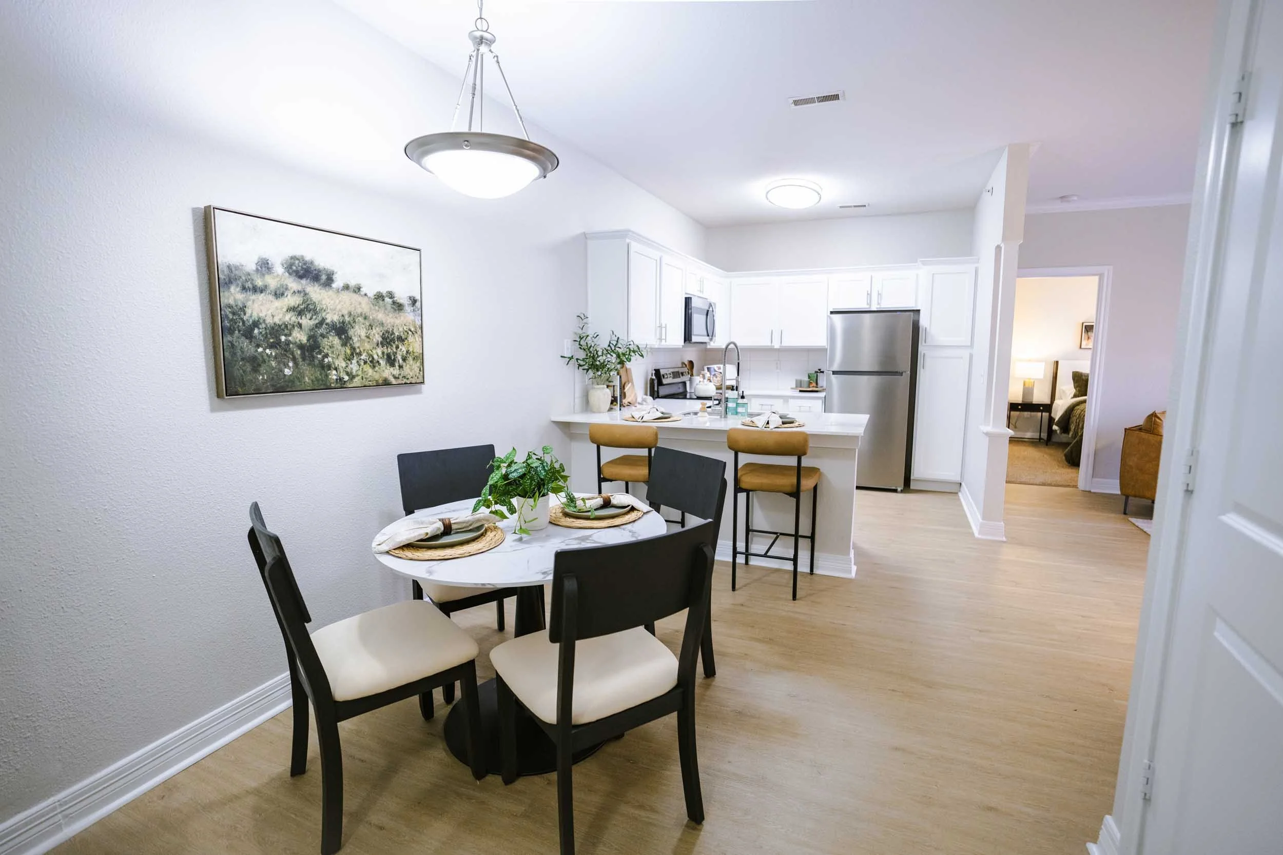 Kitchen and dining area with a round table set for four, black chairs with white cushions, white walls, hardwood flooring, a painting on the wall, stainless steel refrigerator, white cabinets, and a bedroom visible through a doorway.