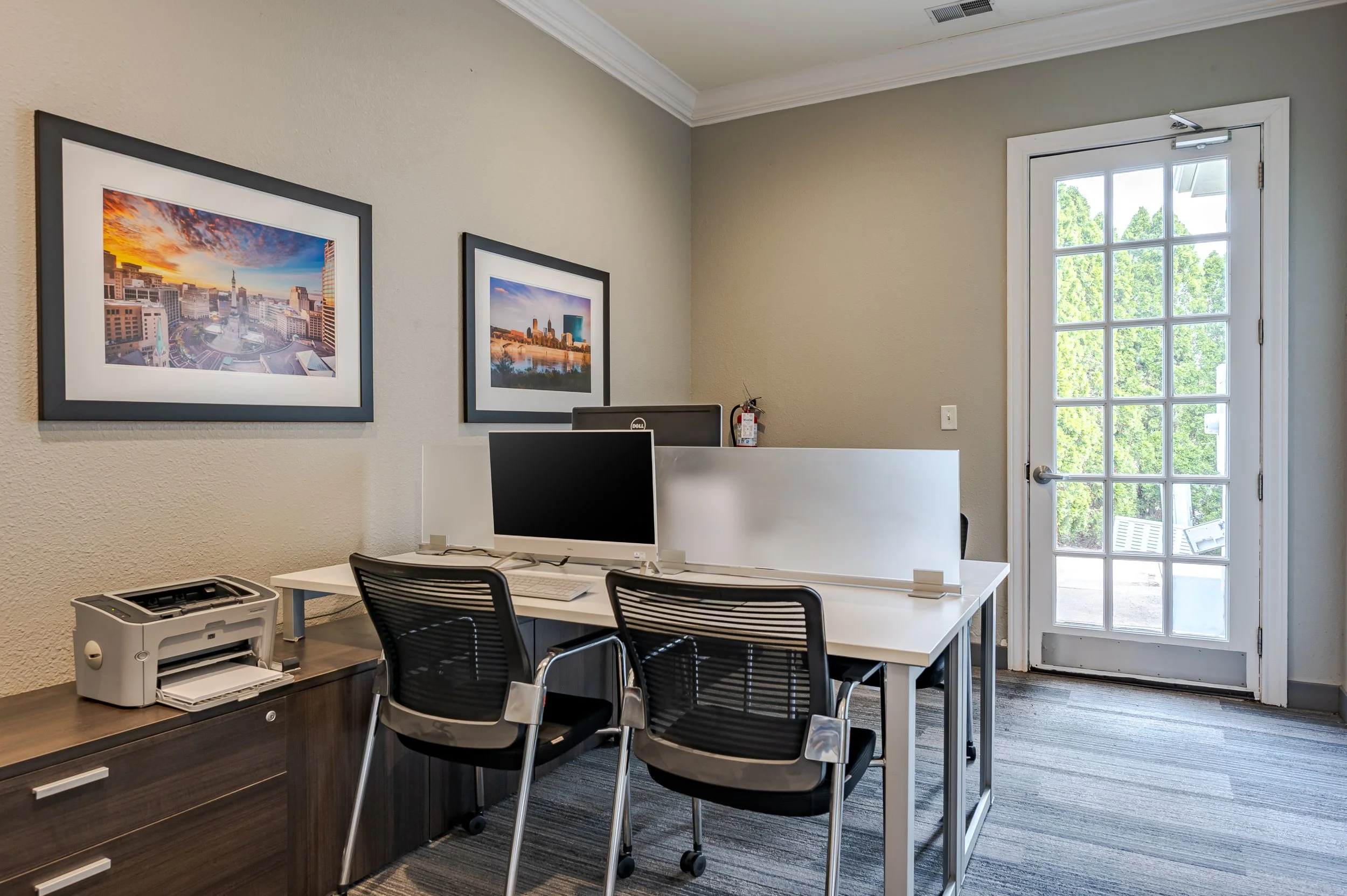 An office with two framed cityscape pictures on the beige wall, a white desk with two black chairs, a computer monitor, a printer, and a fire extinguisher near the door with glass panels showing green trees outside.