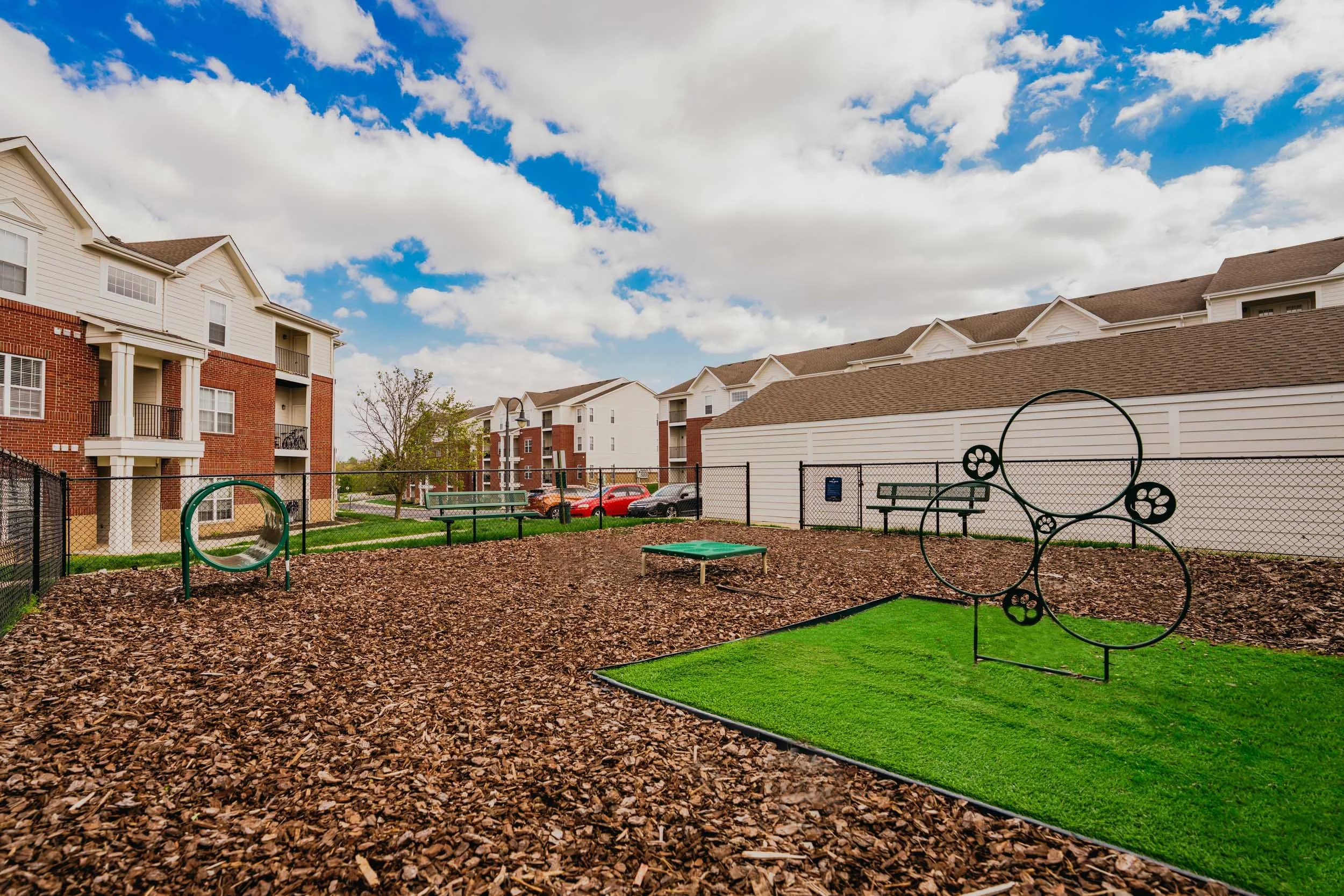 A playground with artificial green turf and wood chip ground cover, surrounded by apartment buildings and a parking lot with cars, under a partly cloudy sky.