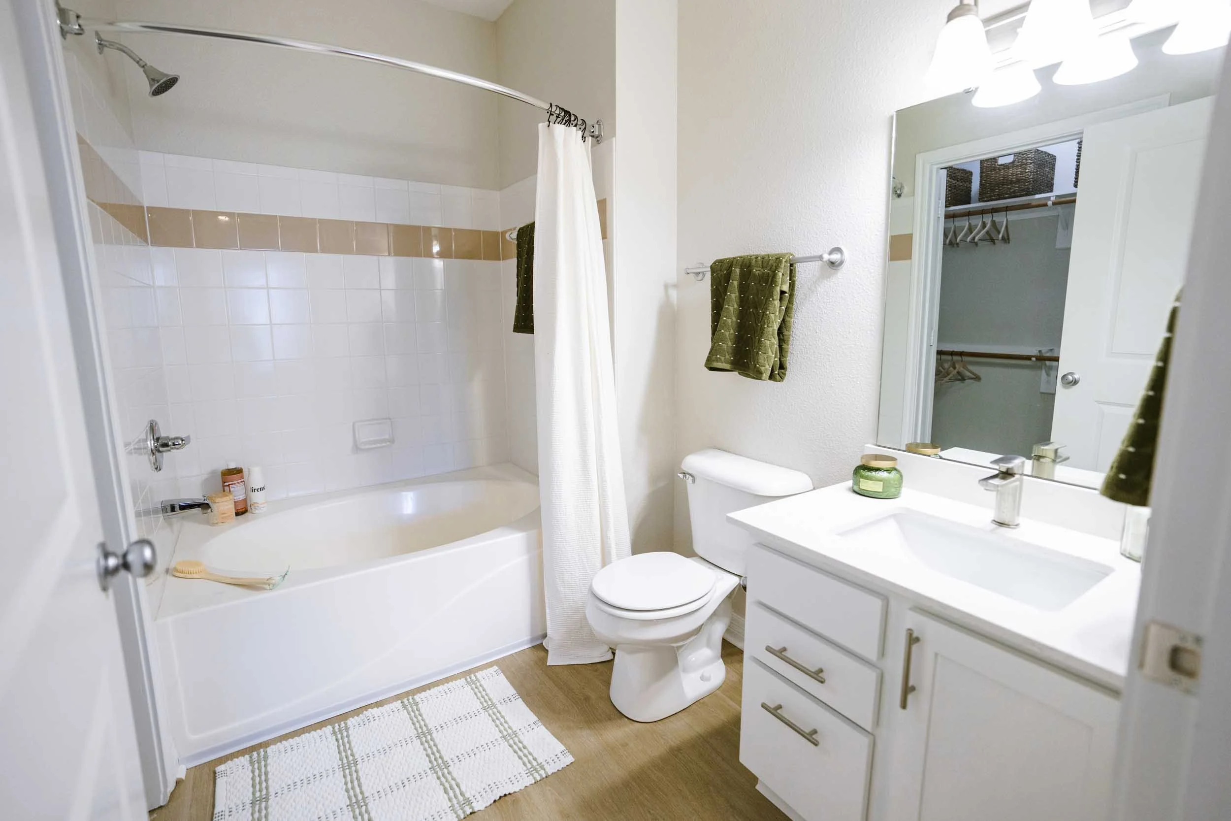 A bathroom with a white vanity, a mirror, a toilet, and a shower with a curtain. Towel racks with green towels, toiletries on the bathtub, and a closet with hangers are visible.