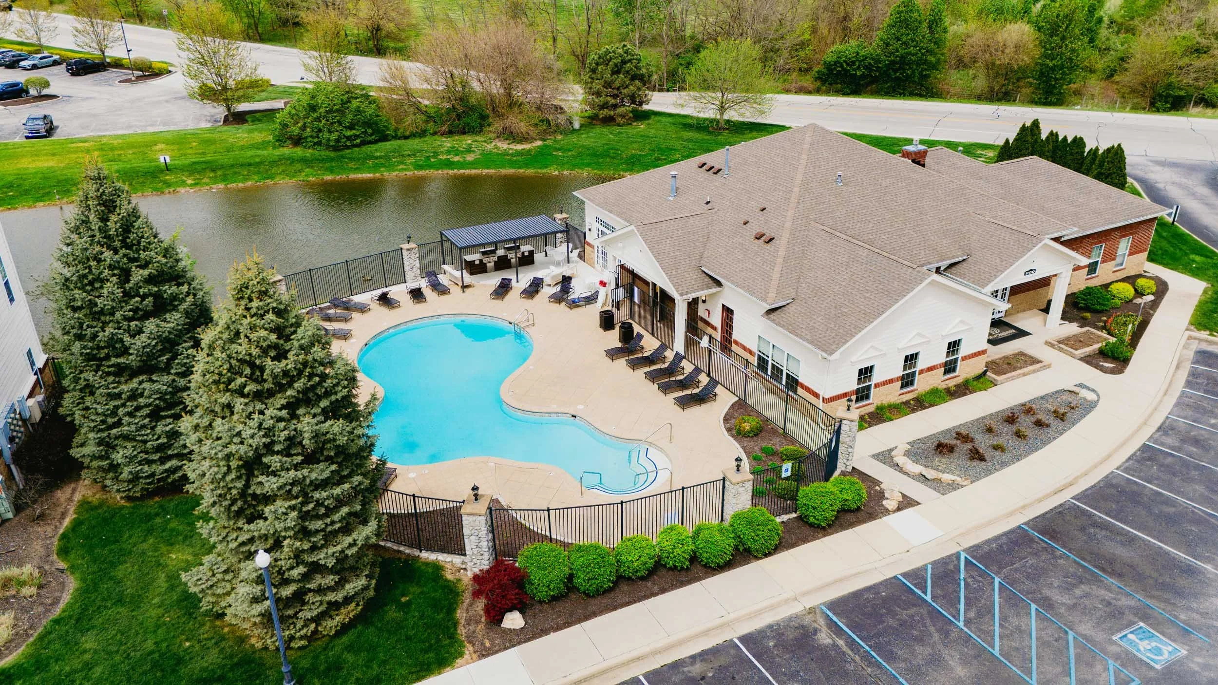 Aerial view of a residential apartment complex with a swimming pool, surrounded by trees, grassy areas, a parking lot with marked spaces, and a small pond behind the building.