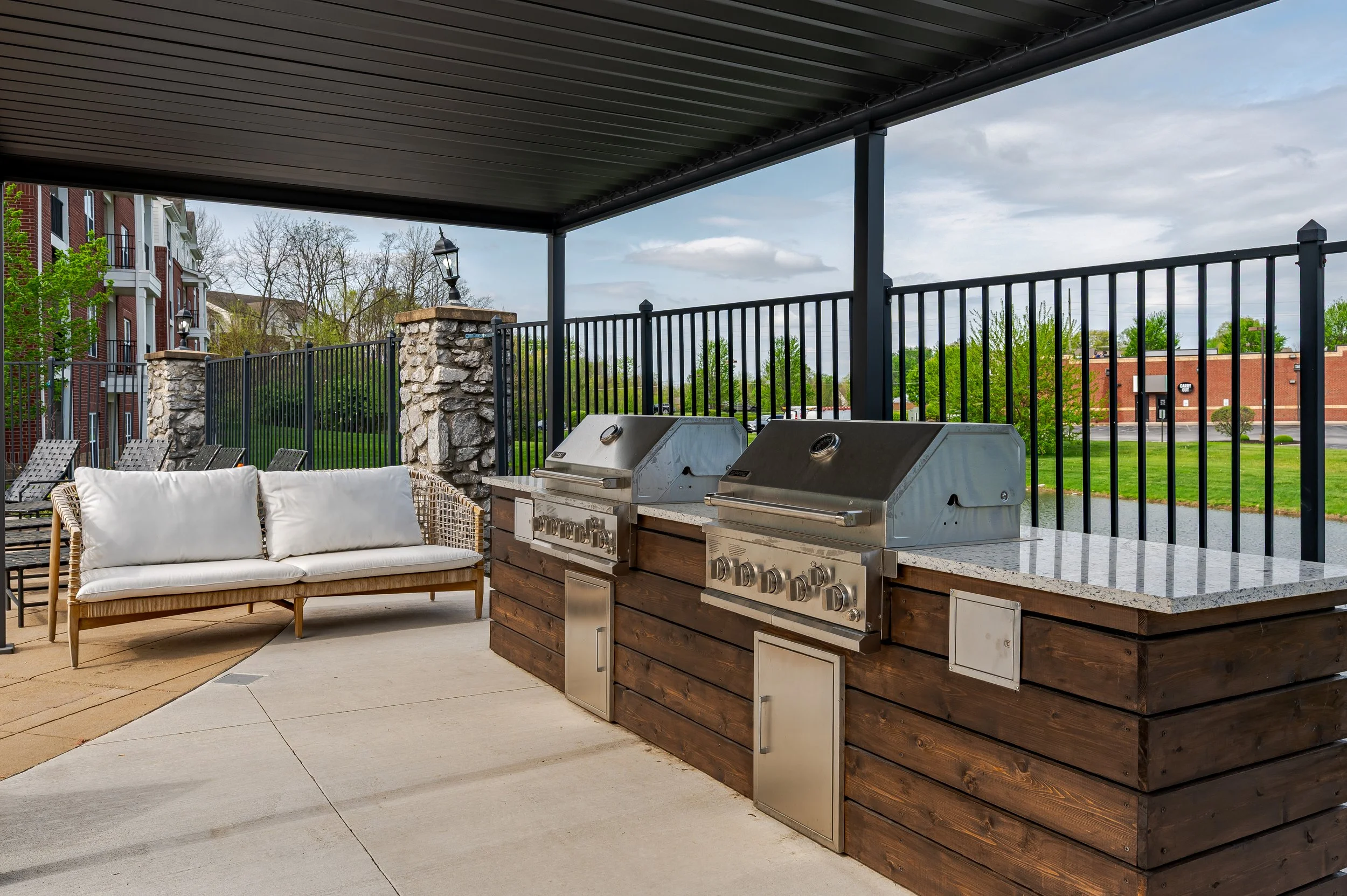 Outdoor patio area with two stainless steel grills, a wicker sofa with white cushions, a stone and wood fire pit table, and a black metal railing under a dark metal roof. There are buildings and trees in the background.
