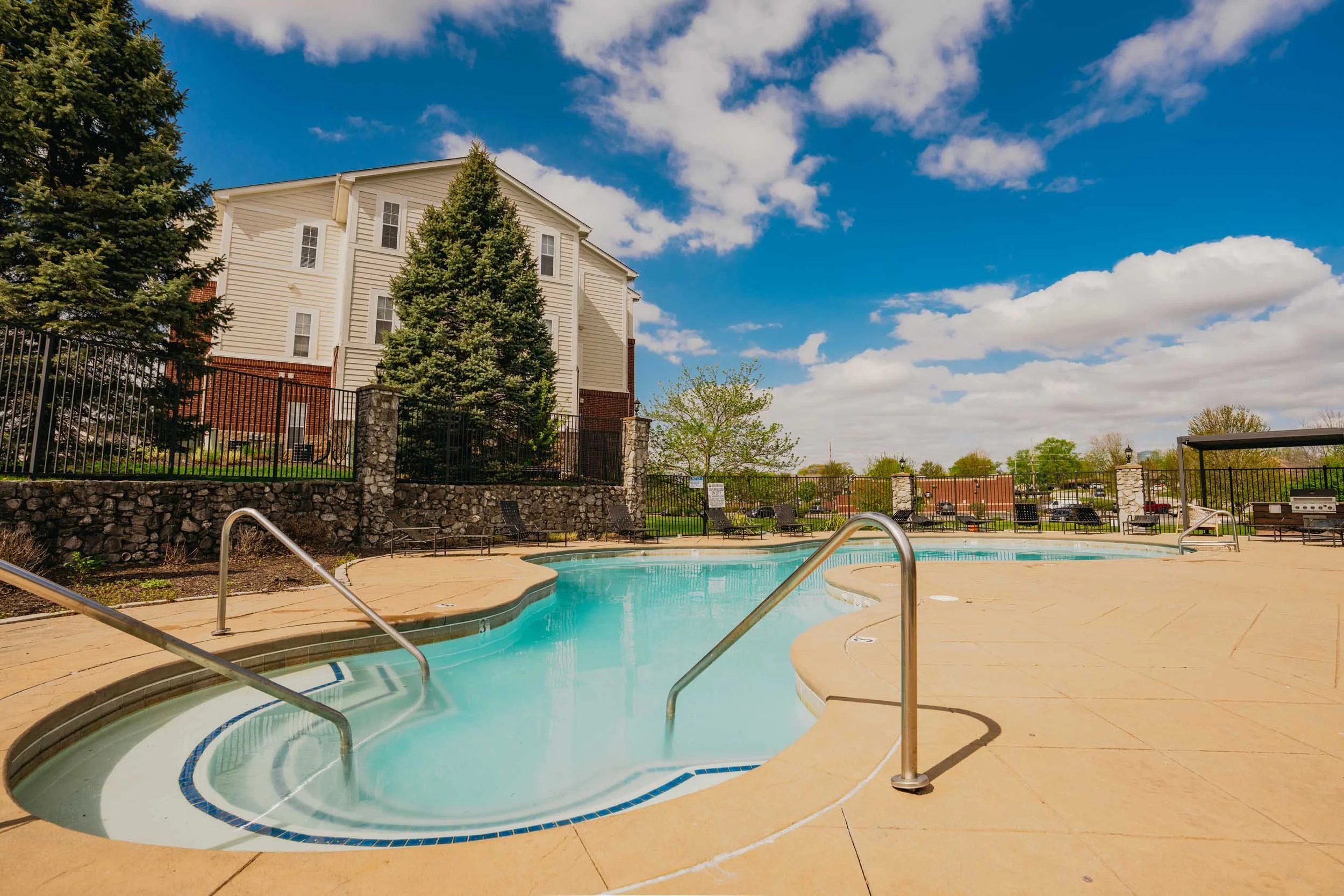 An outdoor swimming pool with a metal handrail and steps, surrounded by a concrete deck and lounge chairs. Behind the pool, there's a stone wall with a black metal fence, tall green trees, and a multistory building with beige siding and red bricks. The sky is partly cloudy with patches of blue.