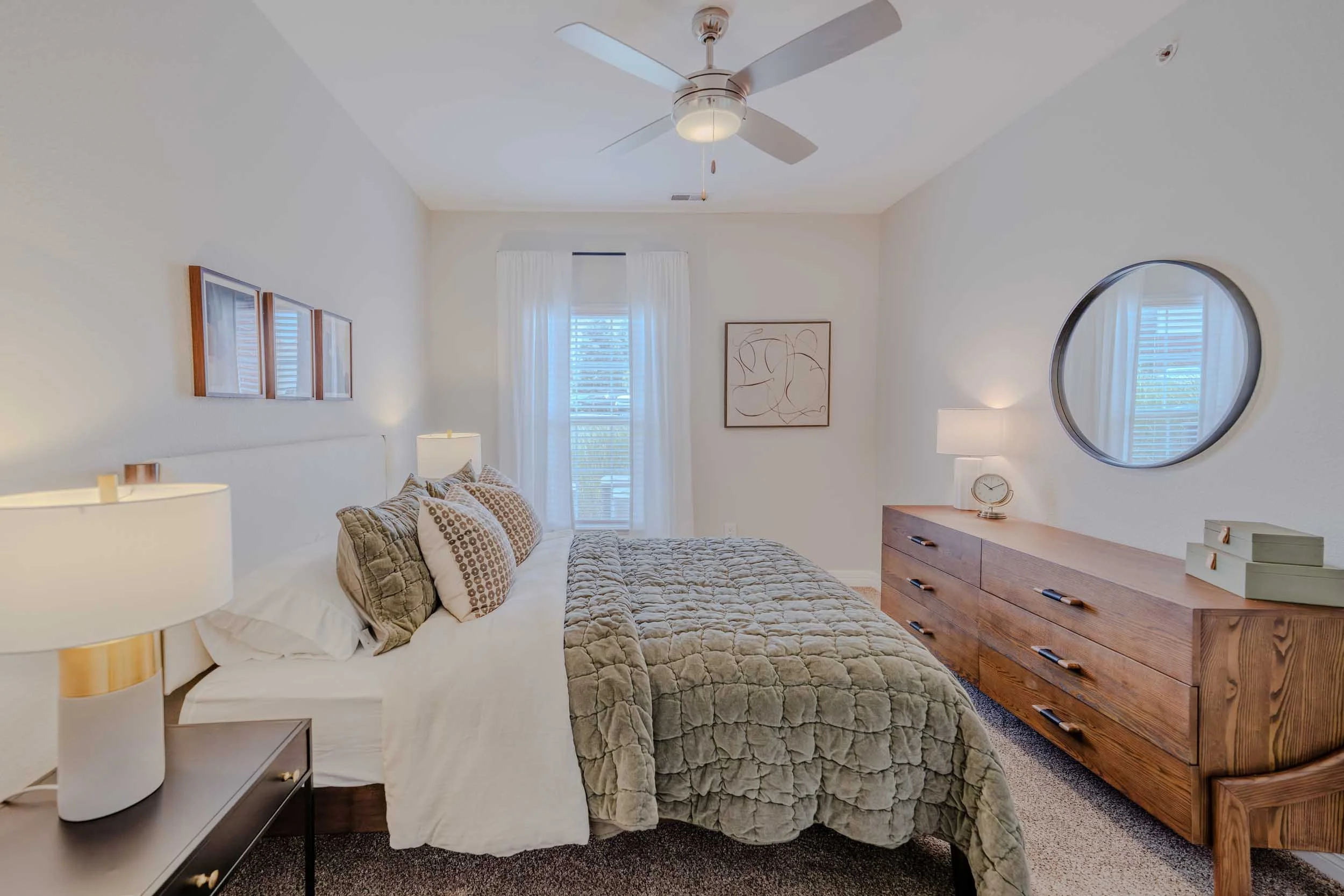 A bedroom with a bed, bedside table, lamps, and wall decor, illuminated with natural light from windows and a ceiling fan.