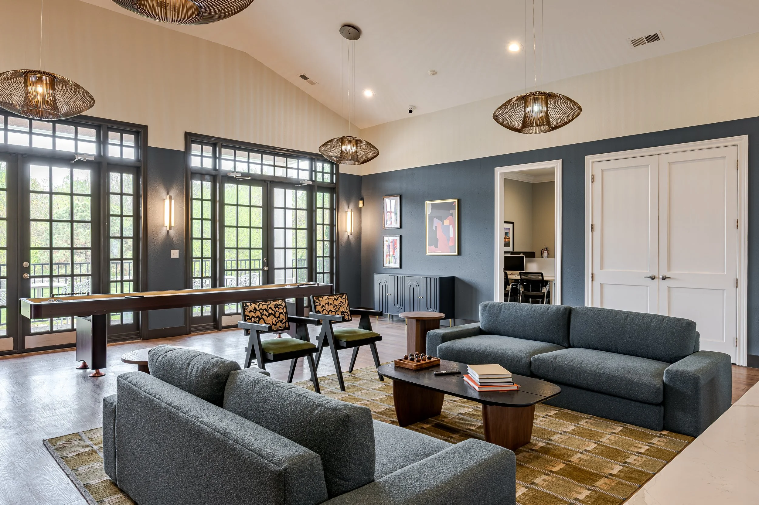 A spacious modern living room with large black framed glass doors and windows, dark blue and white walls, and contemporary furniture including gray couches and wooden chairs. There are modern light fixtures, artwork on the wall, and a wooden coffee t