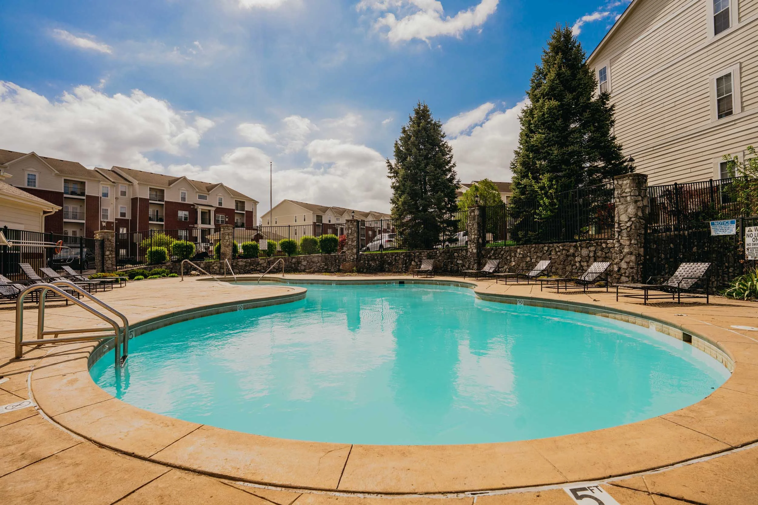 A swimming pool surrounded by lounge chairs, black metal fence, houses, and tall trees with a blue sky and clouds overhead.