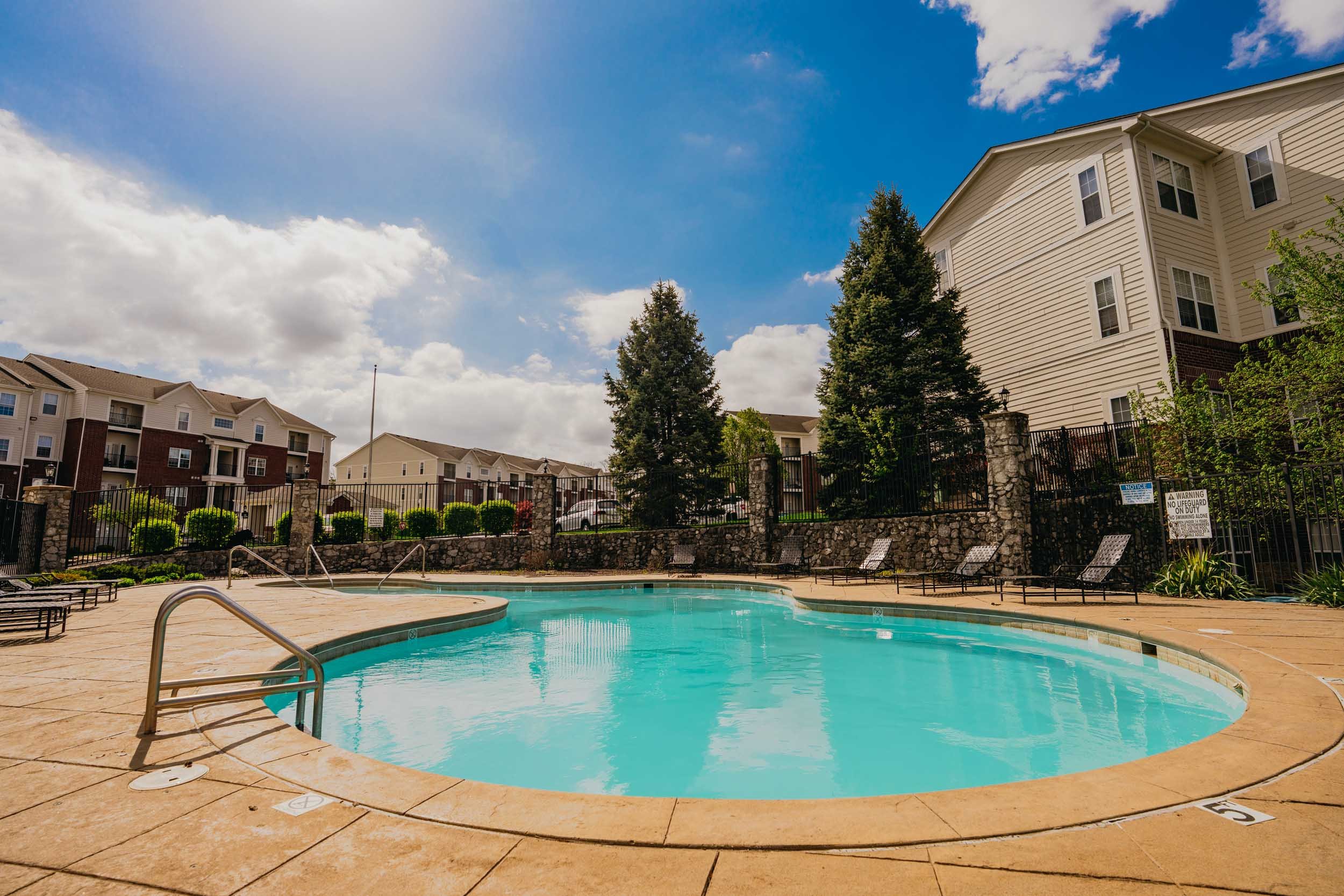 Outdoor swimming pool with lounge chairs, surrounded by a stone fence, tall trees, and residential buildings in the background under a blue sky with clouds.