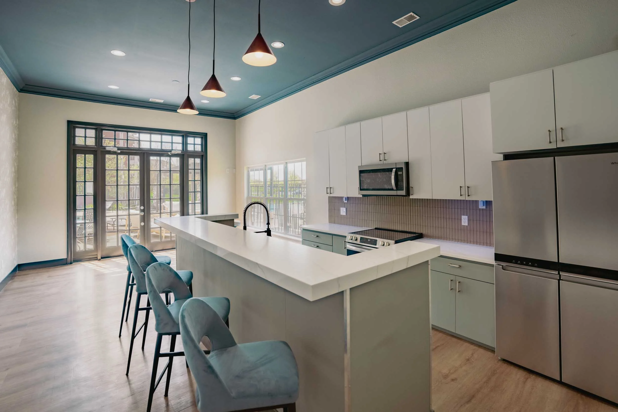 Modern kitchen with white cabinets, a marble island, a black faucet, and stainless steel appliances including a microwave and refrigerator. There are large windows and a door with glass panes, and blue chairs surround the island. The ceiling has rece