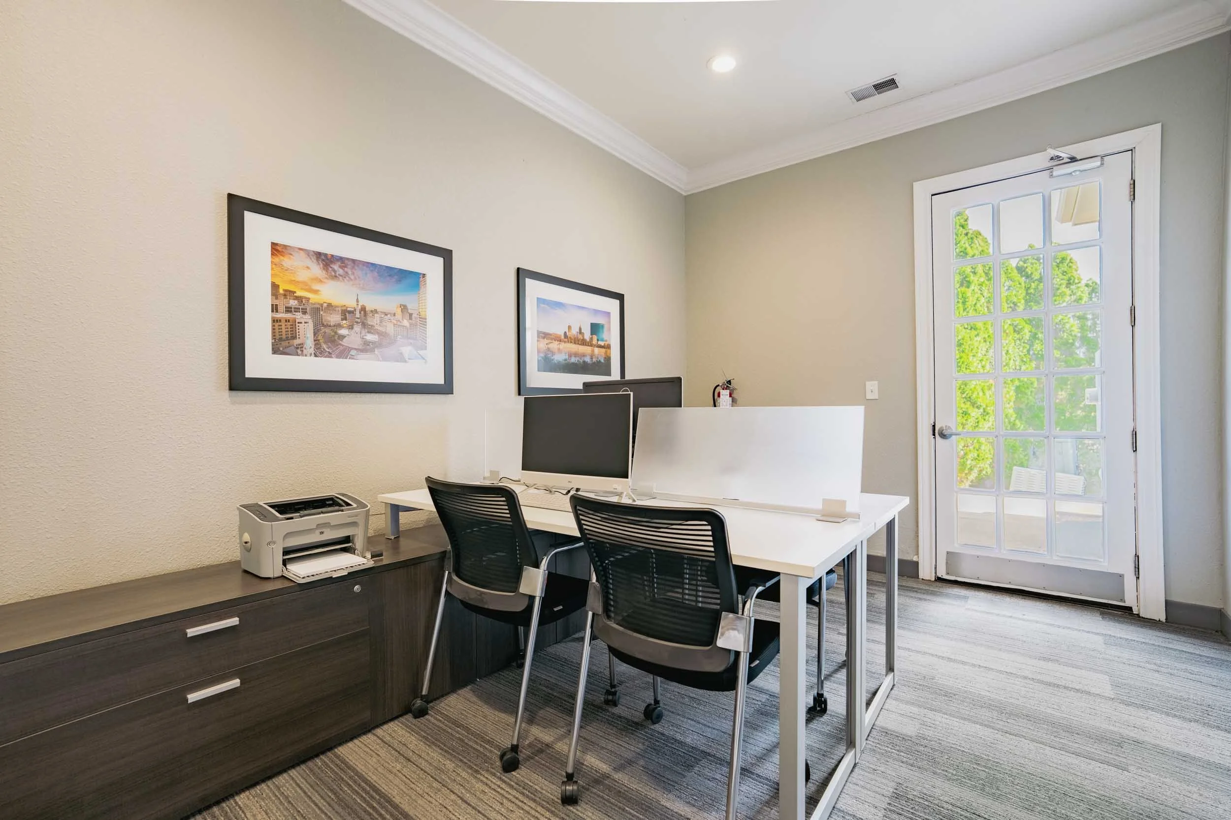 An office with a white desk and two black office chairs, a computer monitor, a printer, and framed cityscape photographs hanging on the beige wall. There is a door with glass panels revealing green trees outside.
