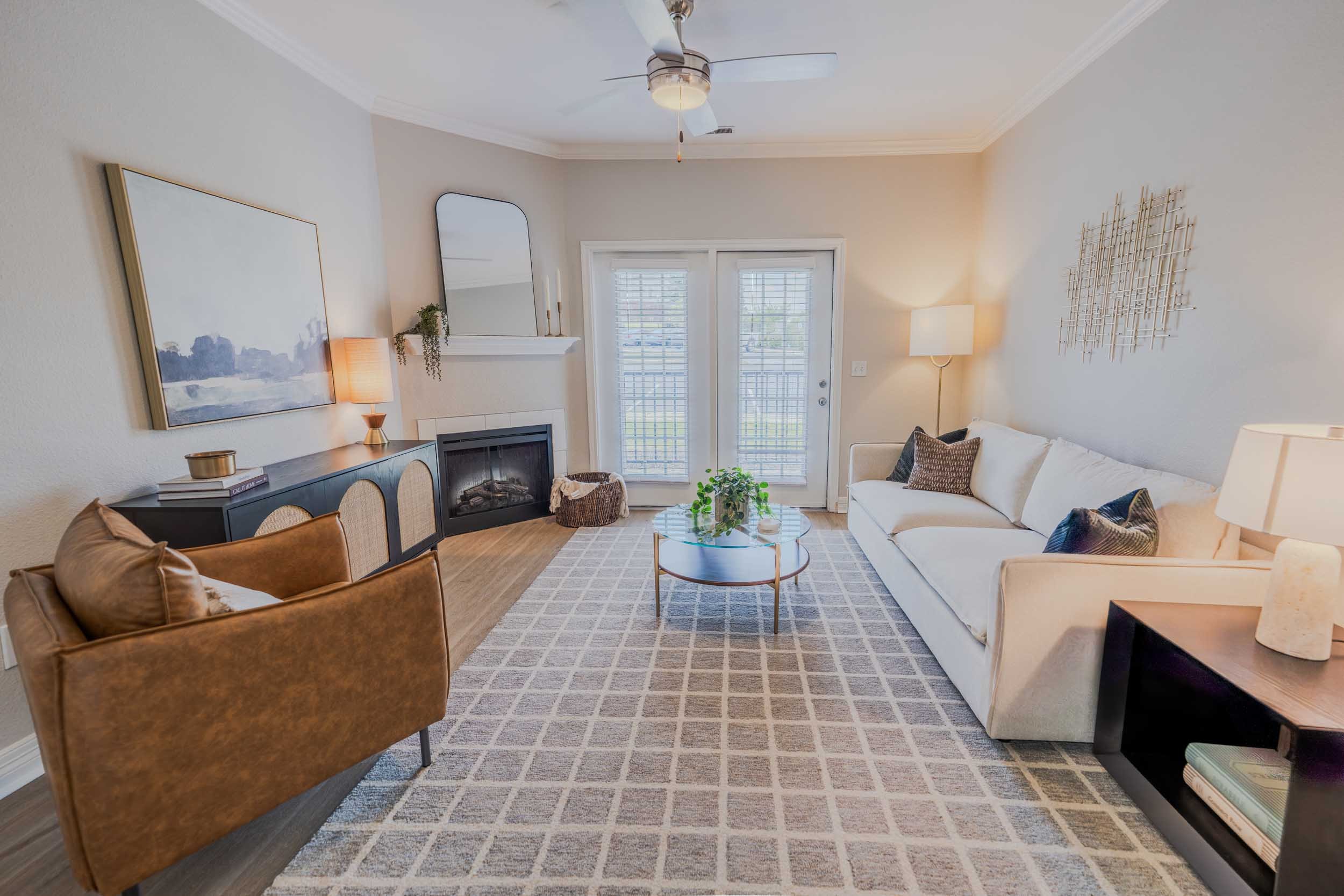 Living room with a white sofa, brown armchair, fireplace, glass coffee table, gray carpet, large windows with blinds, painting above the fireplace, and decor including lamps, plants, and wall art.