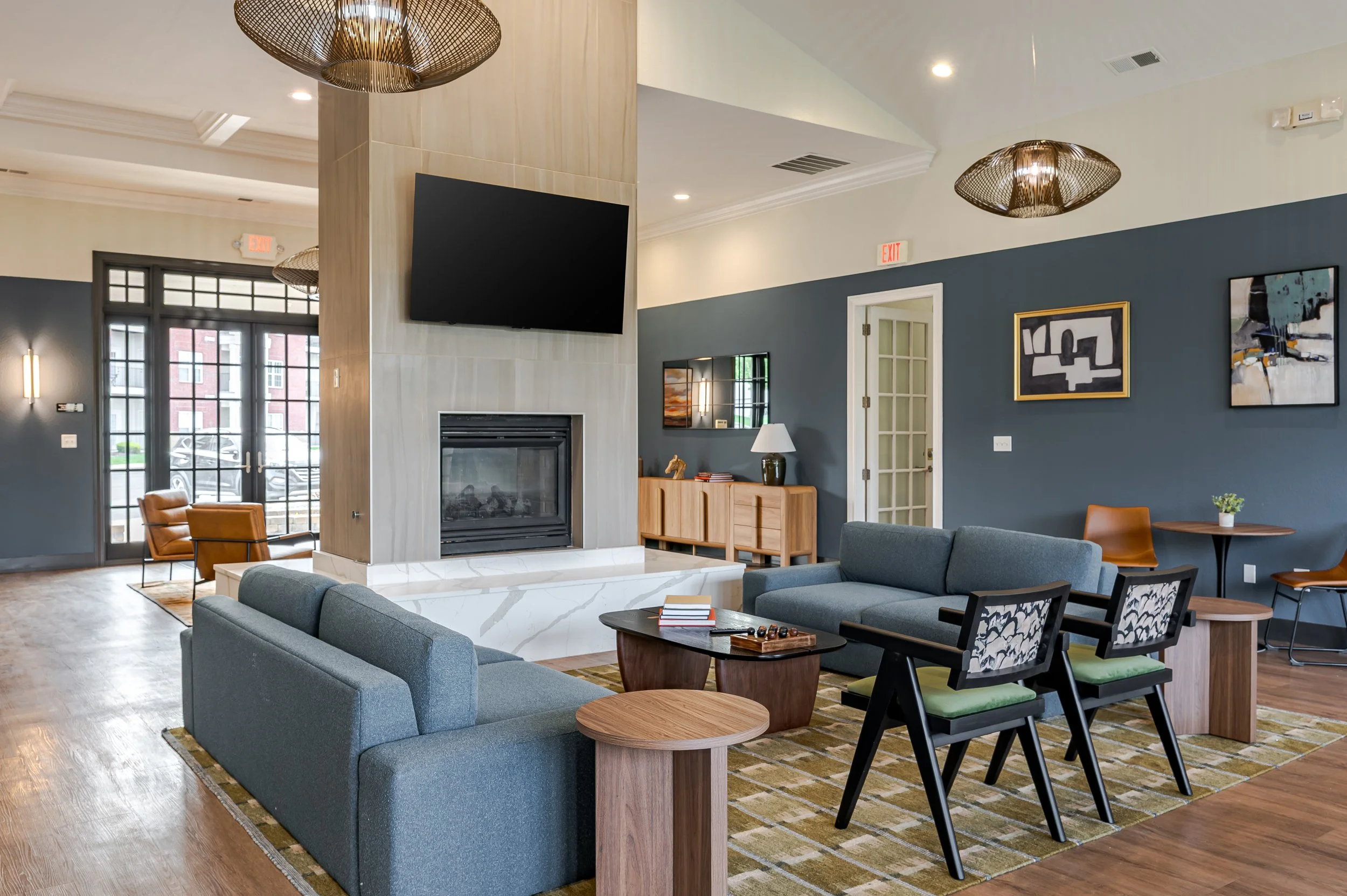 Interior of a modern living room with a fireplace, TV mounted above, blue and neutral colored furniture, artwork on the wall, and large windows/double doors with natural light.