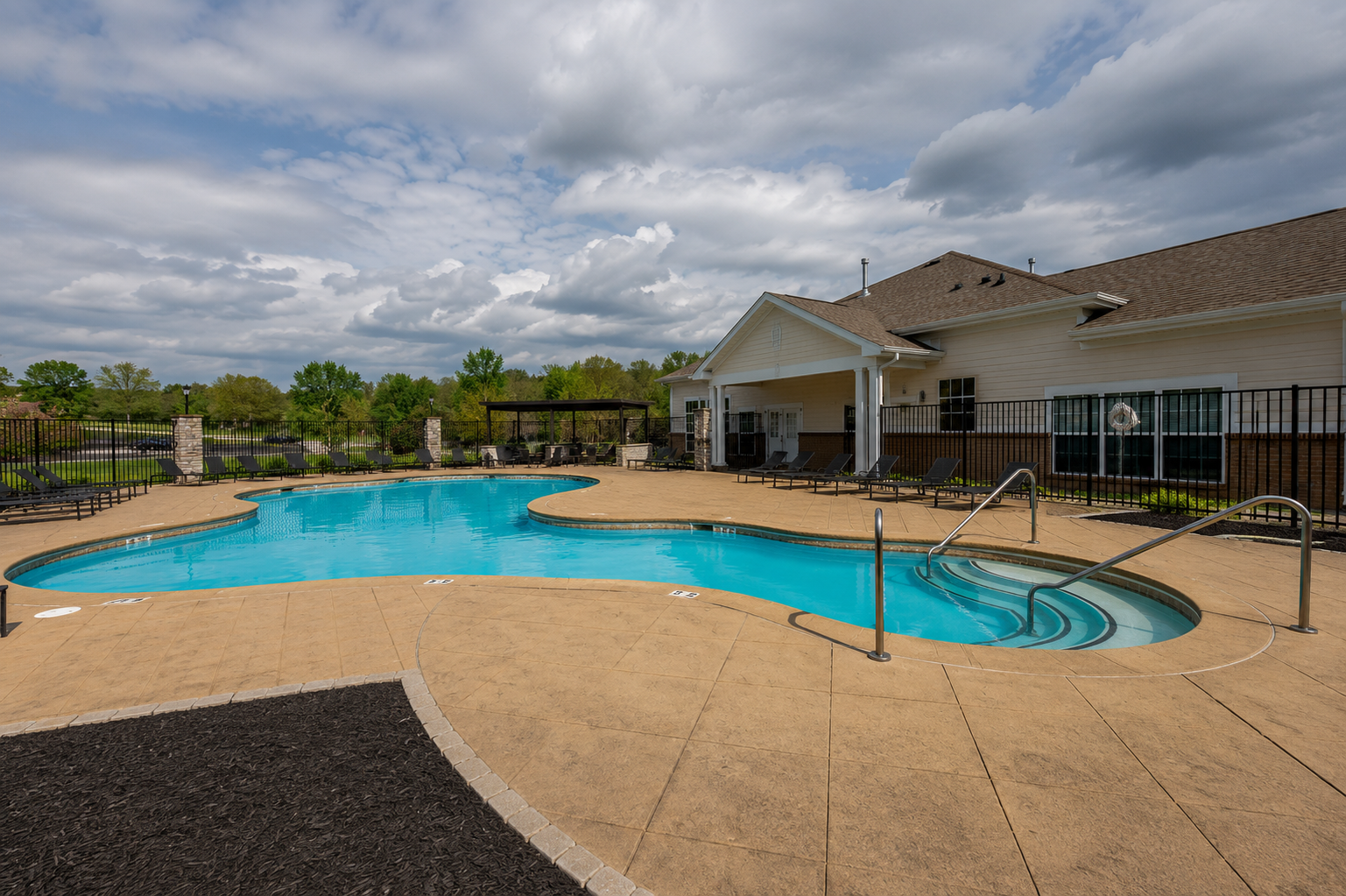 Empty outdoor swimming pool with a tan concrete deck, surrounded by black metal fencing, in a residential area with green trees and cloudy sky in the background.