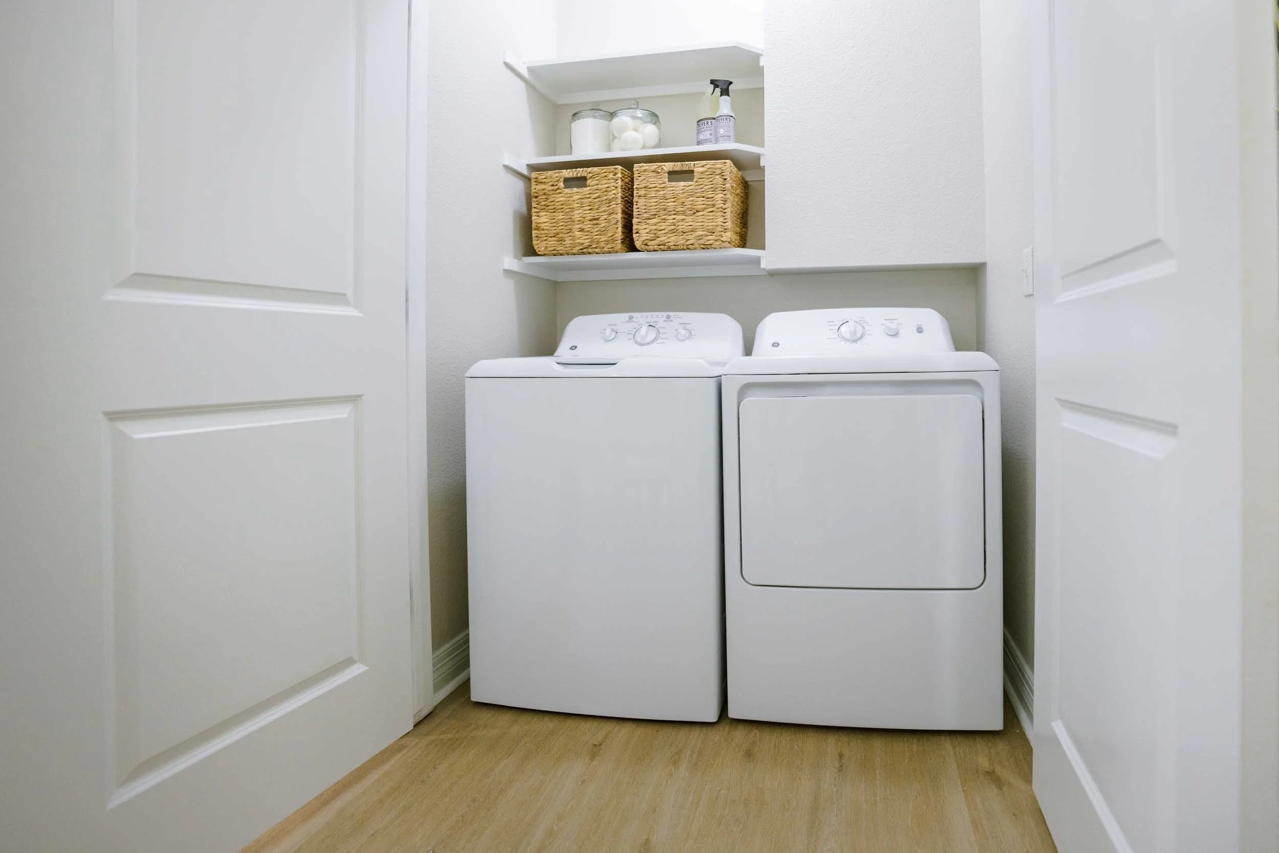 A laundry room with white washer and dryer, two wicker baskets on a white shelf, and cleaning supplies on the top shelf, framed by white cabinet doors.
