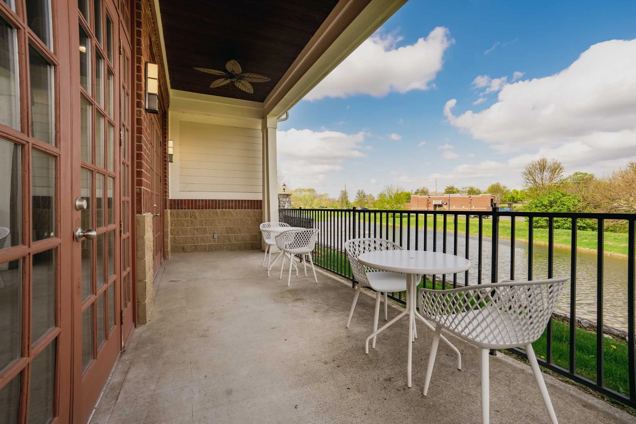 Balcony with white chairs and a round table overlooking a river and trees under a blue sky with some clouds.