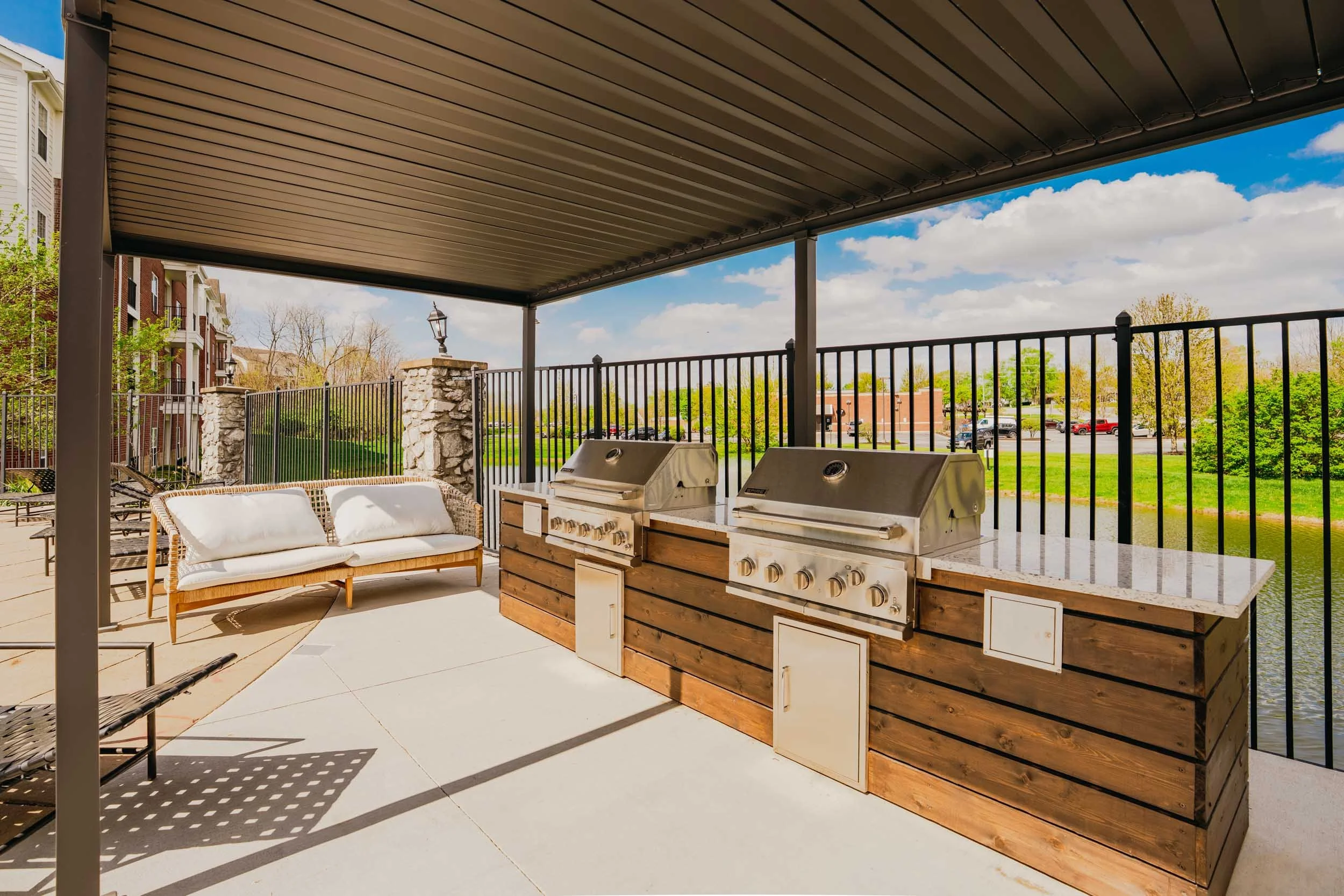 Outdoor porch area with a covered ceiling, a white outdoor sofa, a barbecue grill setup with two grills, and a scenic view of trees, buildings, and a parking lot in the background.