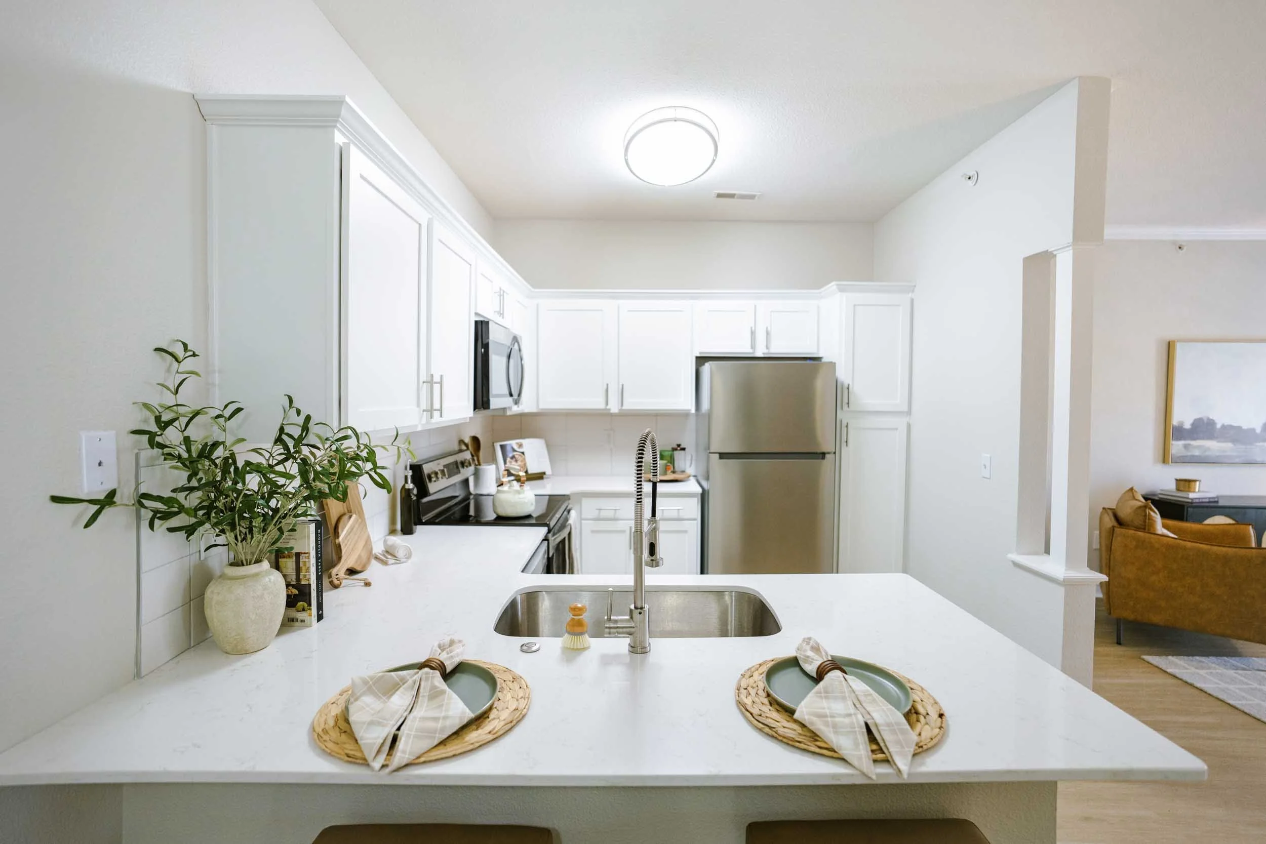 A modern kitchen with white cabinets, a stainless steel refrigerator, a stove, and a microwave. There is a white countertop with a plant and kitchen items, and a kitchen island with place settings for two.