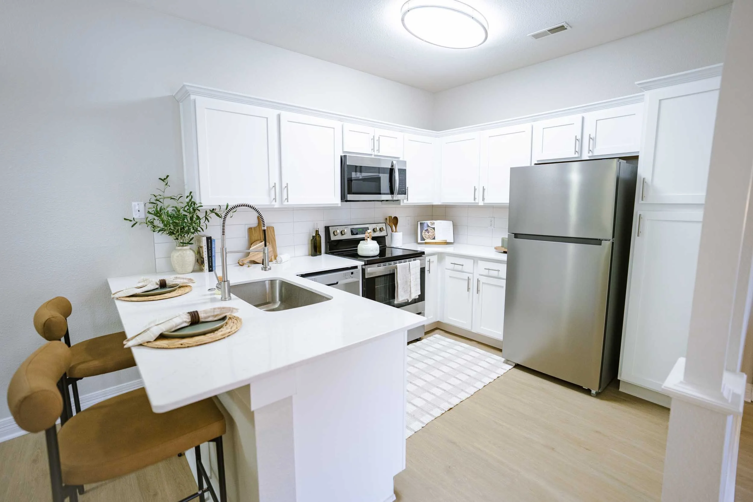 A modern kitchen with white cabinets, stainless steel appliances, a kitchen island with a sink, and a small dining area with three brown chairs and place settings. There is a potted plant on the counter near the sink.