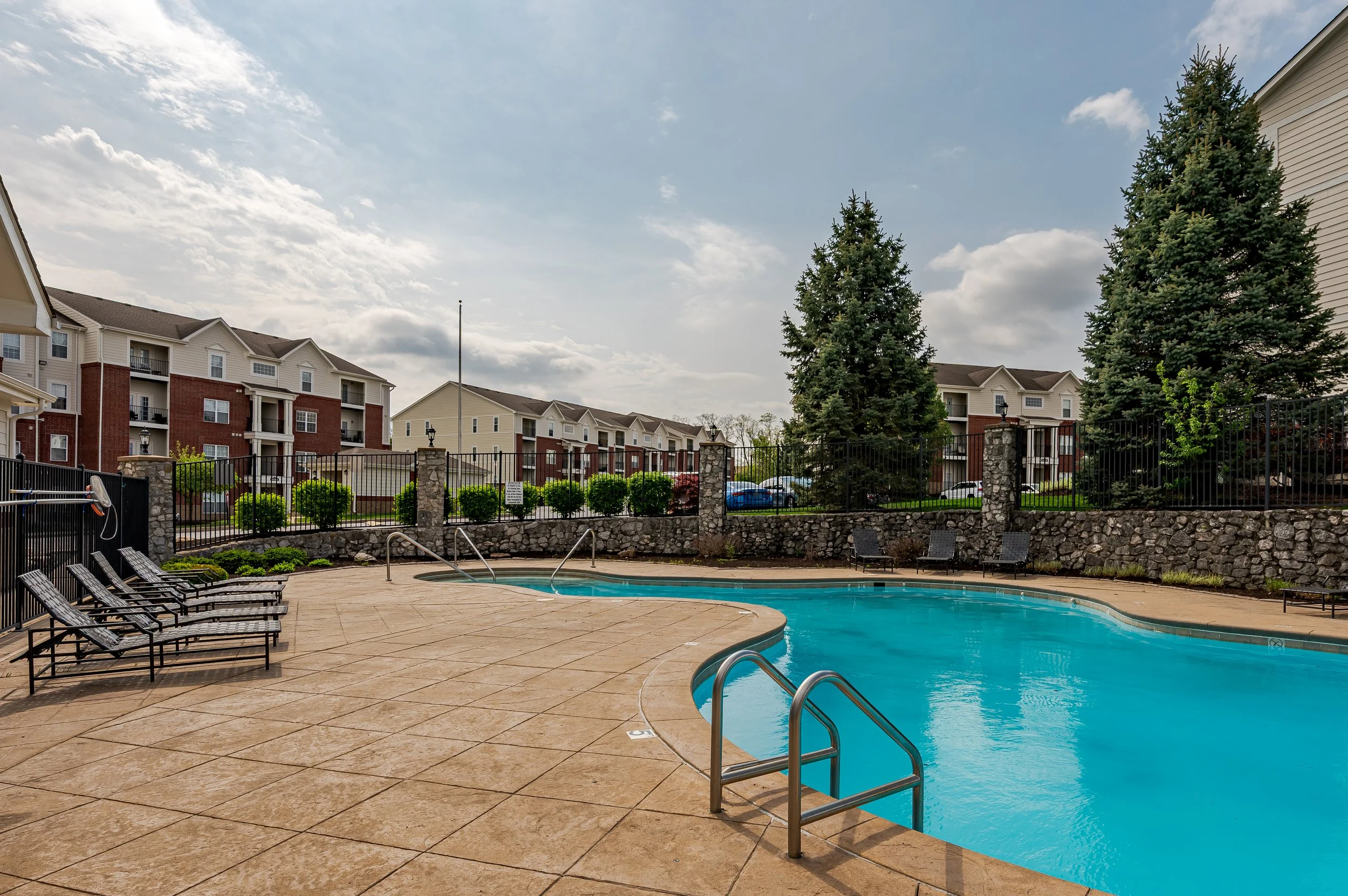 Outdoor swimming pool area surrounded by lounge chairs, backyard with a black metal fence, trees, and residential buildings in the background under a partly cloudy sky.