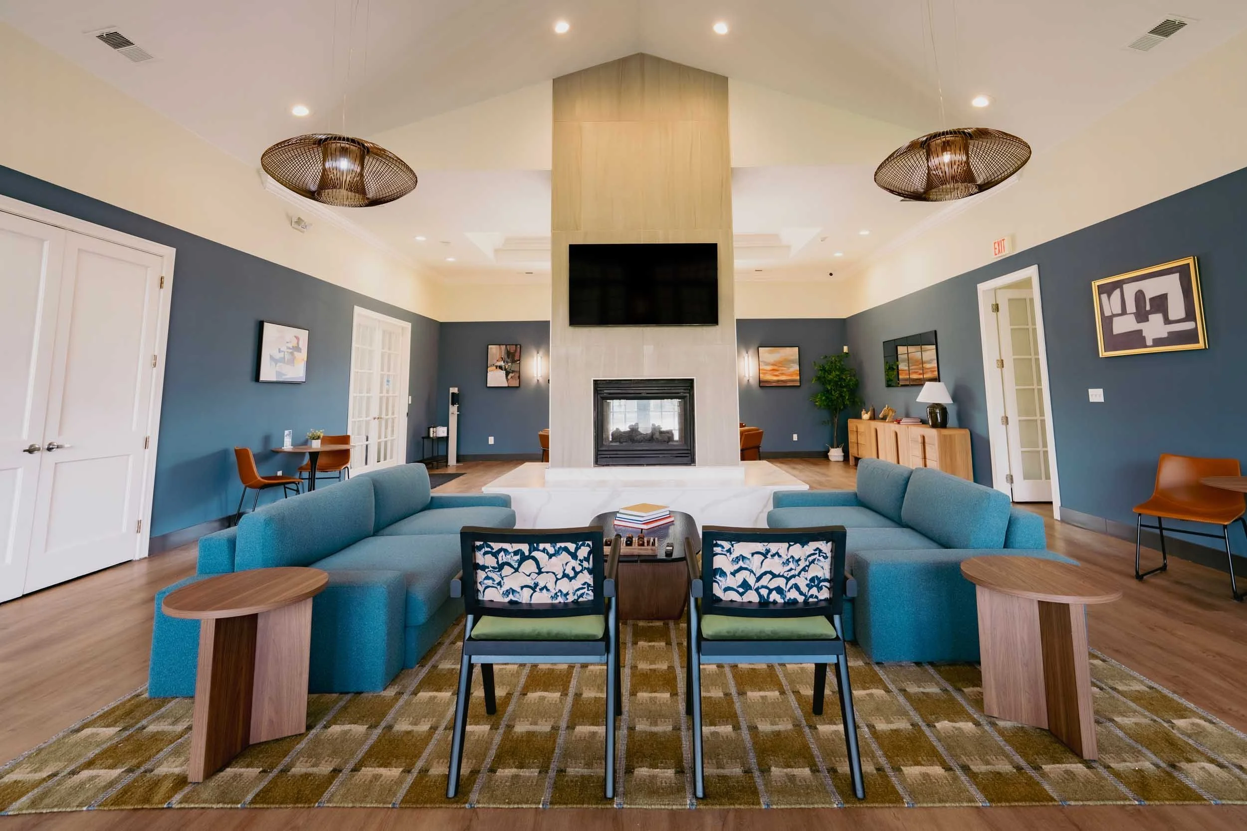 A spacious living room with a high vaulted ceiling, a fireplace, a large flat-screen TV mounted above the fireplace, and comfortable seating including a blue sofa and black-and-white patterned chairs. The room has blue and cream-colored walls with fr