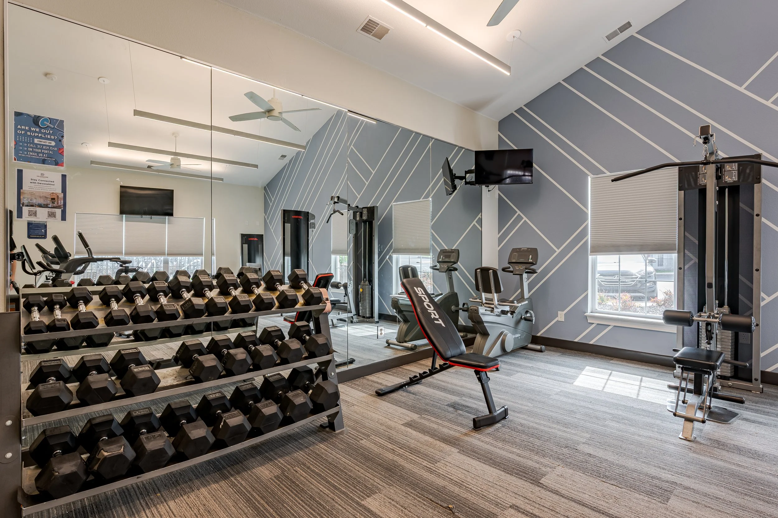 A modern gym with various workout equipment including dumbbells on a rack, cardio machines like an exercise bike, and weight machines. The room has a large mirror on one wall, a blue and white geometric accent wall, windows with blinds, and ceiling f