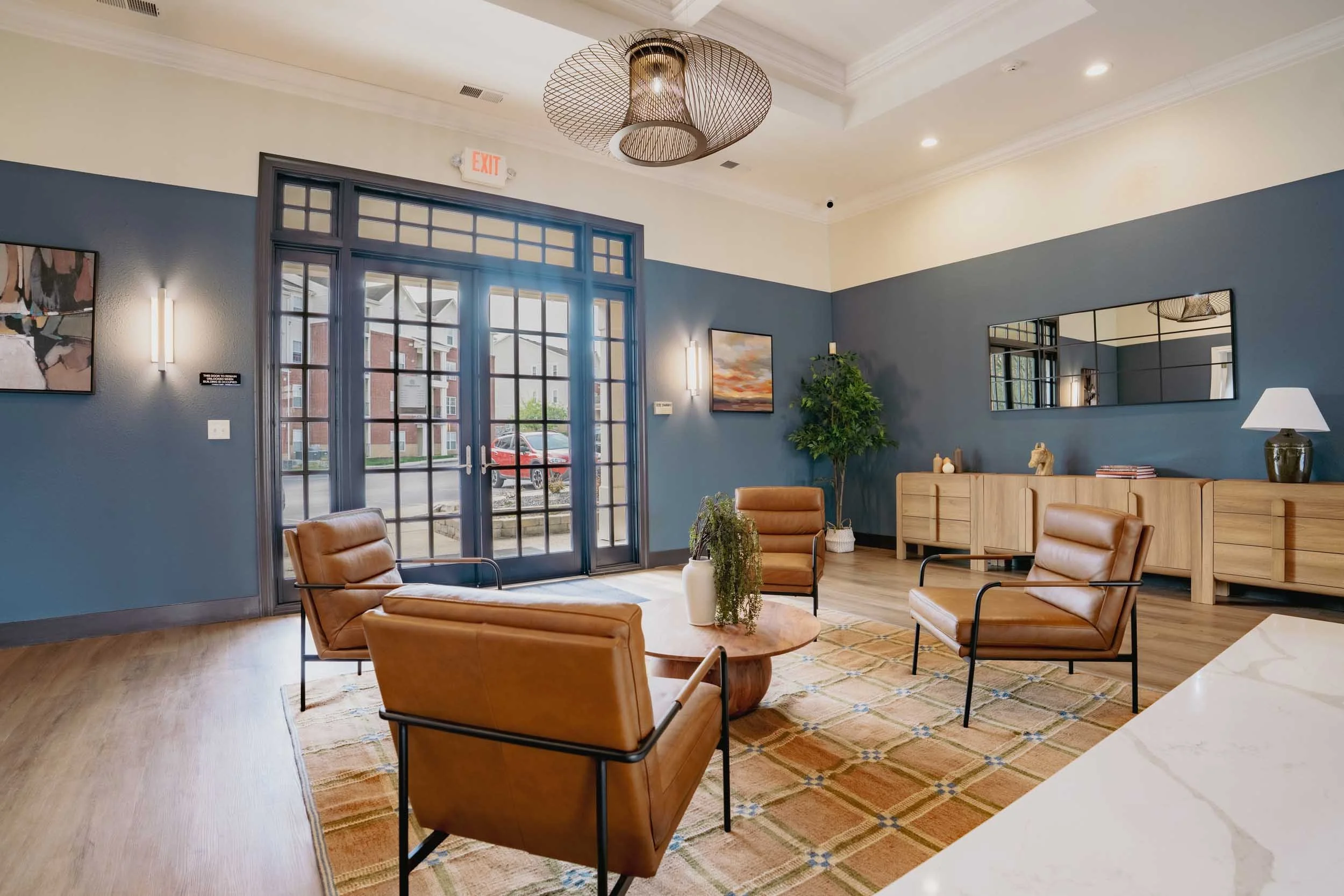 A modern lobby area with leather armchairs, a low wooden table, and a large mirror on a blue wall. There is a potted plant, a lamp, and artwork on the walls. Large glass doors lead outside, letting in natural light. The ceiling has a contemporary lig