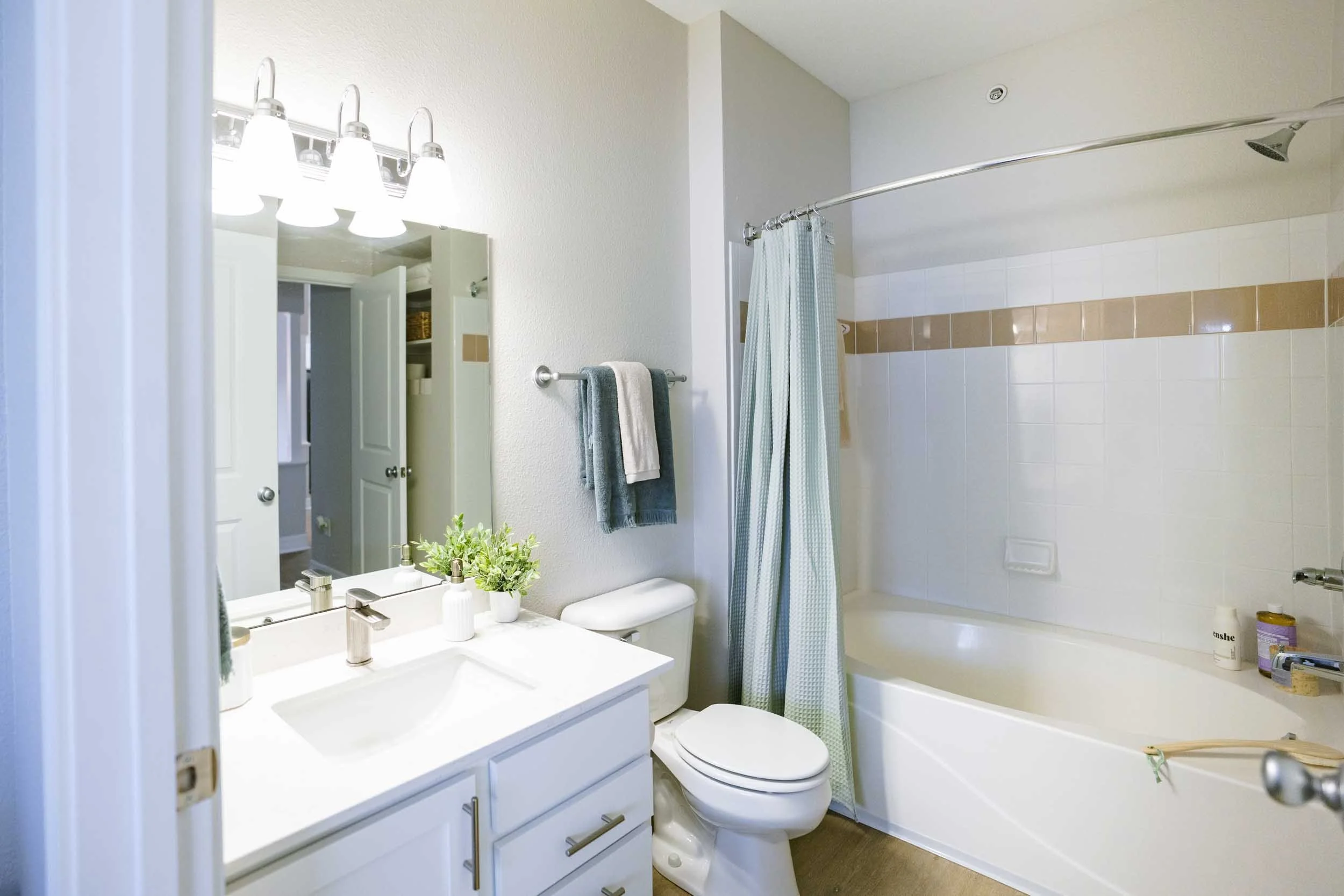 A small bathroom with a white vanity and sink, a mirror with four lights above it, a toilet, and a bathtub with a shower curtain. Decor includes a small plant, towels, and personal care items.