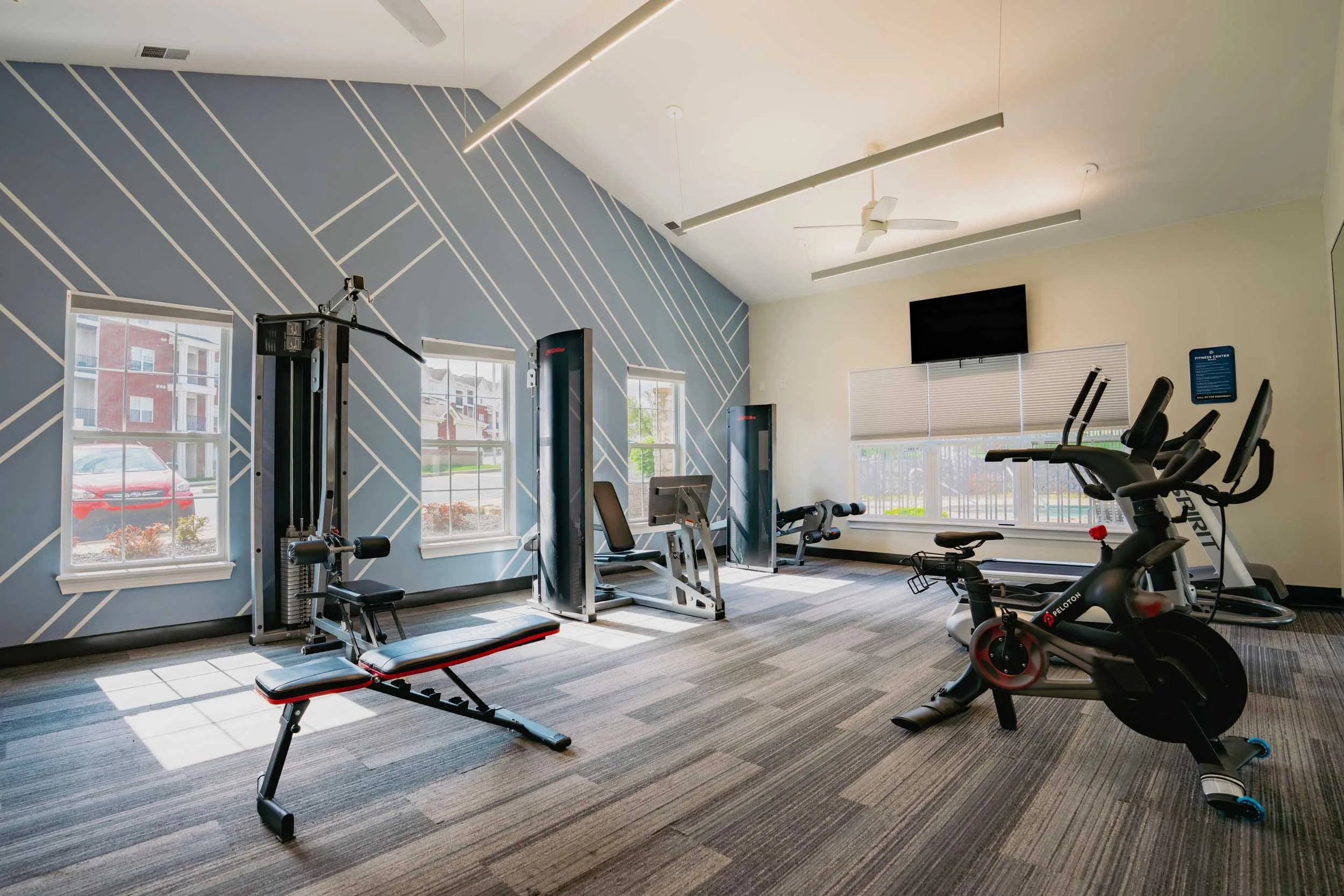 Interior of a fitness center with workout equipment including a weight machine, treadmill, stationary bike, and elliptical trainer. Large windows let in natural light, and there is a wall-mounted television and ceiling fans.