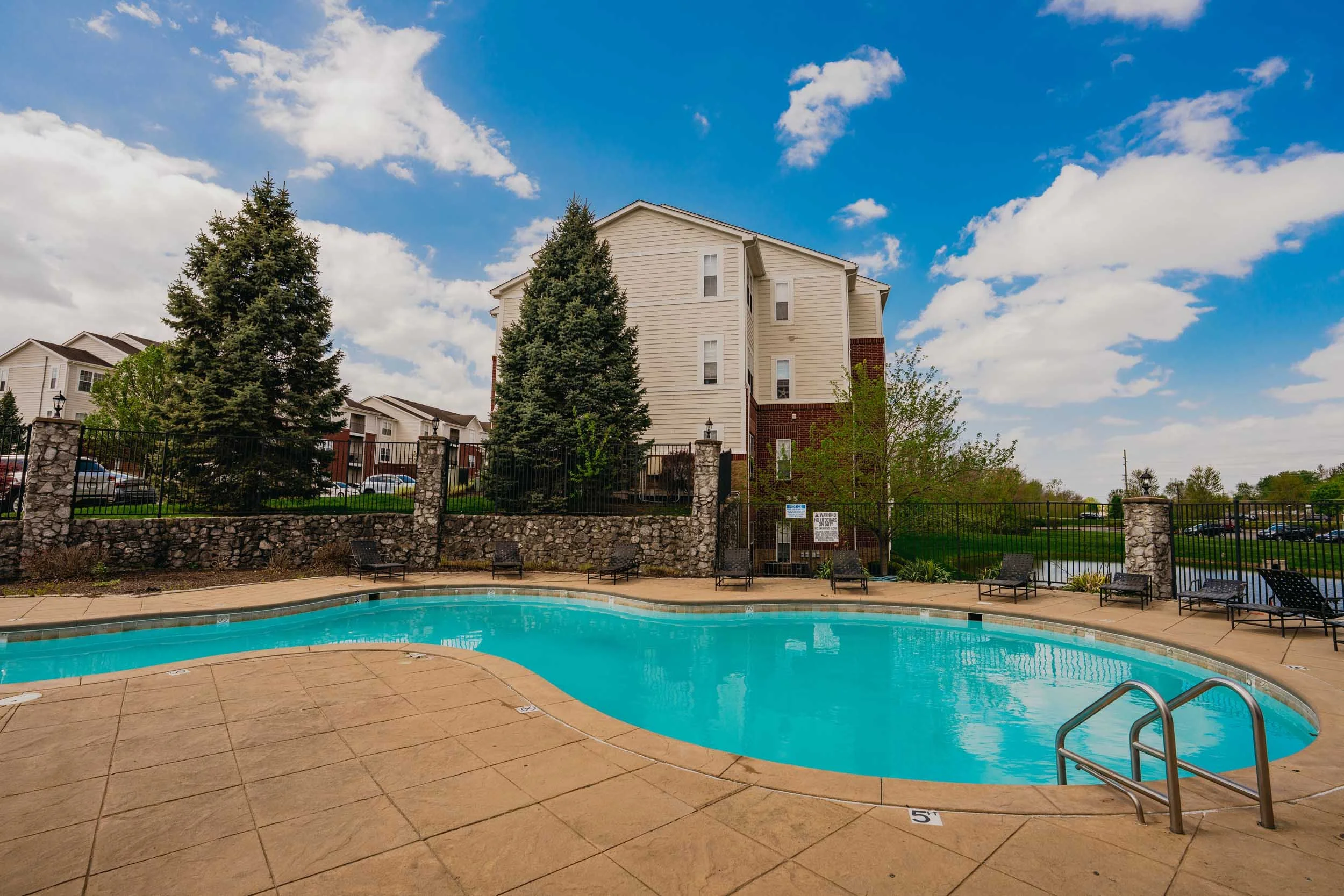 Outdoor swimming pool with a beige tiled deck, surrounded by a black metal fence, in front of apartment buildings and tall evergreen trees under a partly cloudy blue sky.