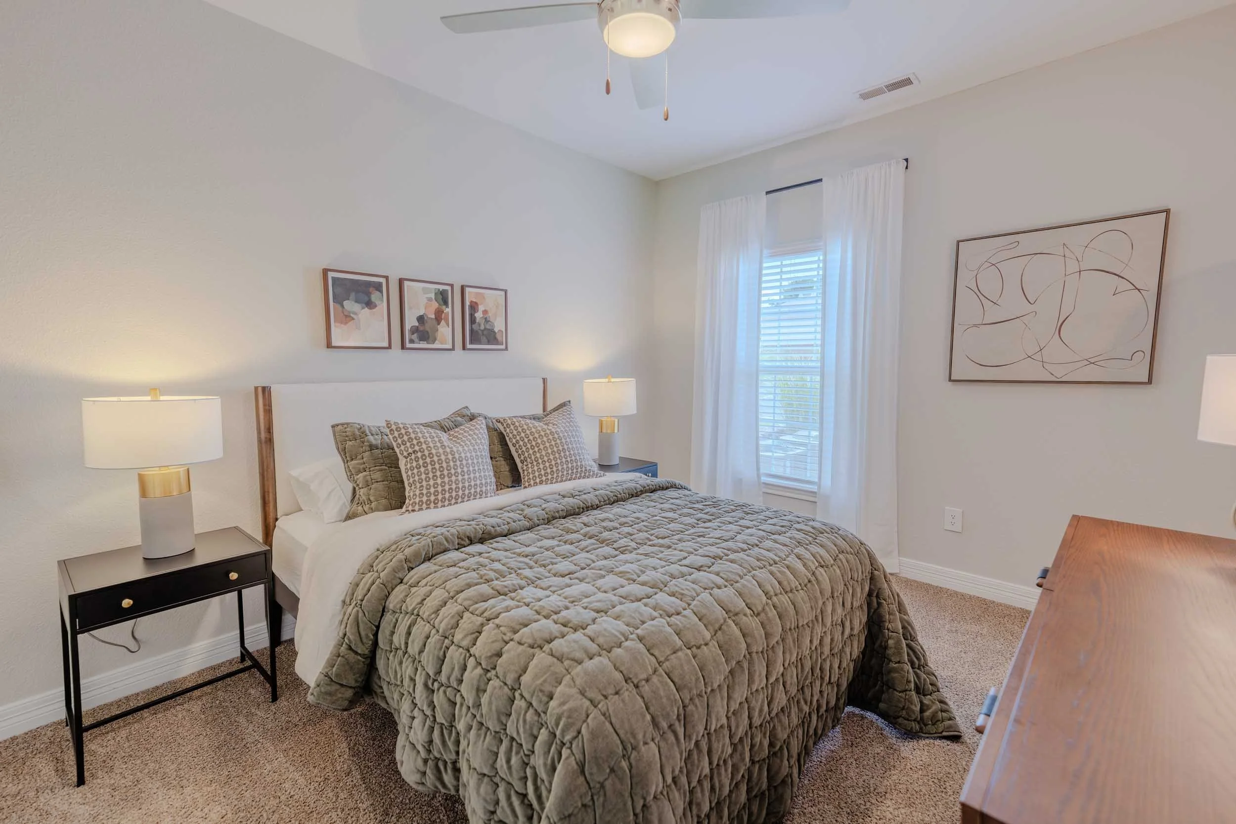A bedroom with a bed featuring a beige quilted comforter and several pillows, two white bedside lamps on dark nightstands, a window with white curtains, three abstract art pieces above the headboard, a ceiling fan with a light, and a wooden dresser t