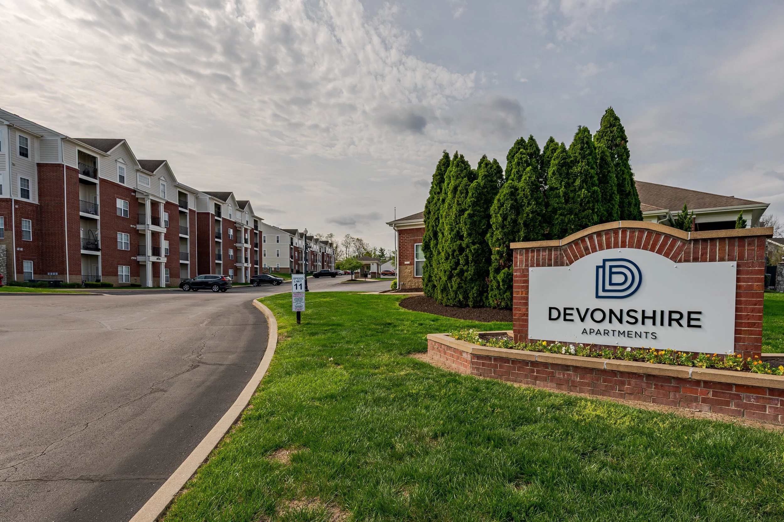 A residential apartment complex with a brick and white siding building, several cars parked on the street, and a sign that reads 'Devonshire Apartments' next to a row of tall green trees under a partly cloudy sky.