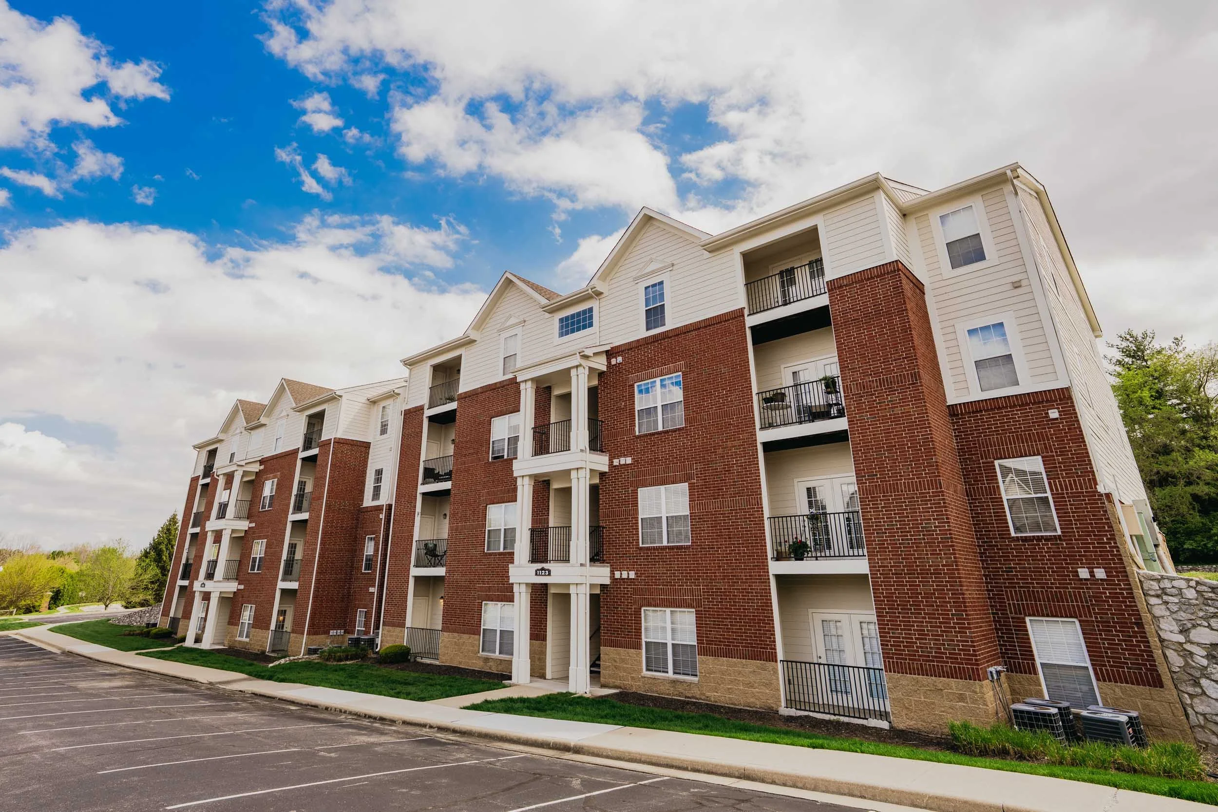 Exterior view of a multi-story residential apartment building with white and brick facade, balconies, and a parking lot in the foreground under a partly cloudy sky.