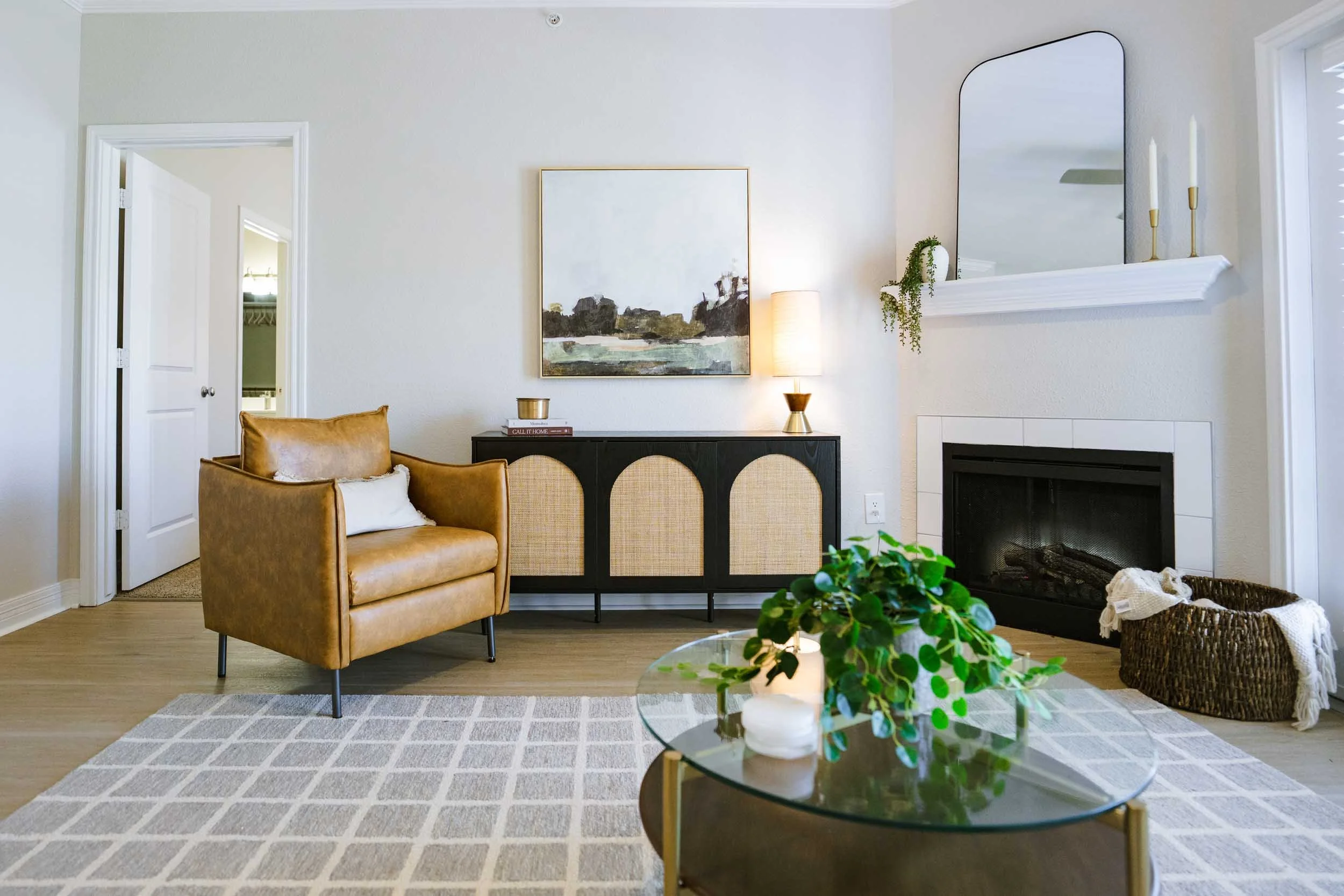 Living room with a tan armchair, a black and rattan sideboard, a white fireplace, a large abstract painting, a tall lamp, a mirror, candles, a potted plant, and a basket of throw blankets.