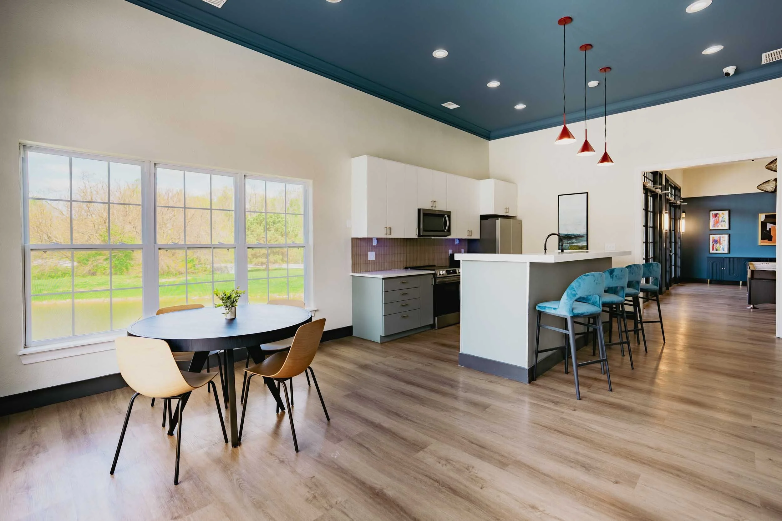 Modern kitchen and dining area with large window, blue ceiling with recessed lighting and pendant lights, white cabinets, grey lower cabinets, stainless steel appliances, and light wood flooring, with a blue accent wall and artwork.