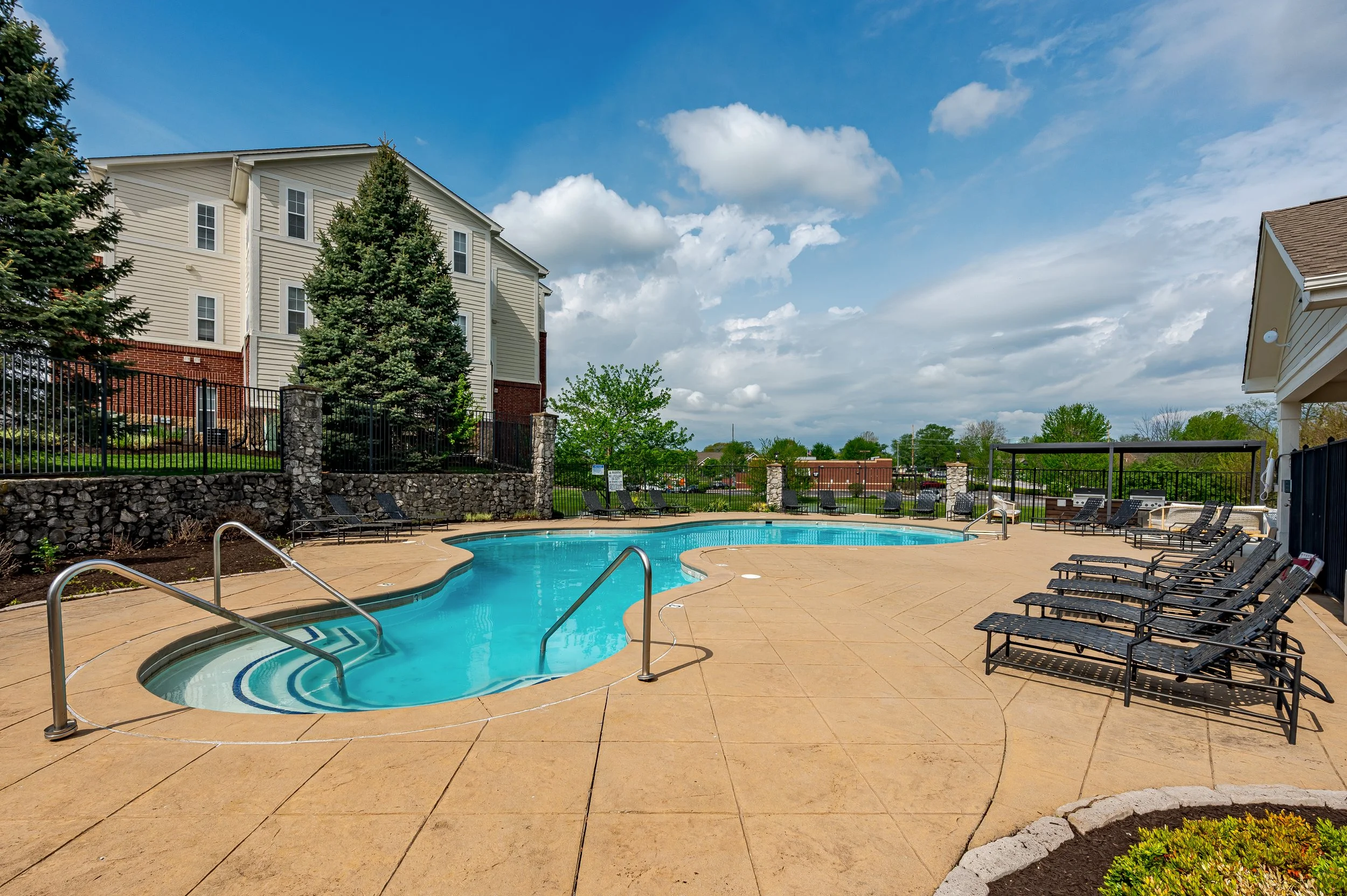 An outdoor swimming pool area in a residential complex with poolside lounge chairs, surrounded by buildings with trees and a partly cloudy sky.