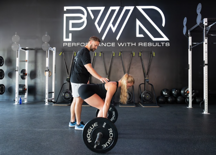A woman lifting a barbell with weight plates while a man supervises her in a gym. The gym has black walls, fitness equipment, and a large logo saying 'BW Performance with Results' on the wall.