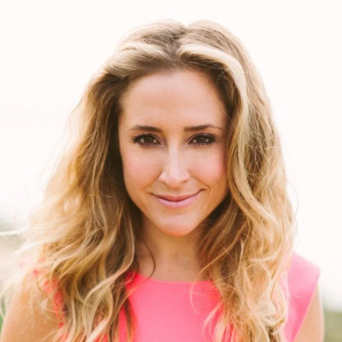 Smiling woman with long wavy blonde hair wearing a pink top against a light background.