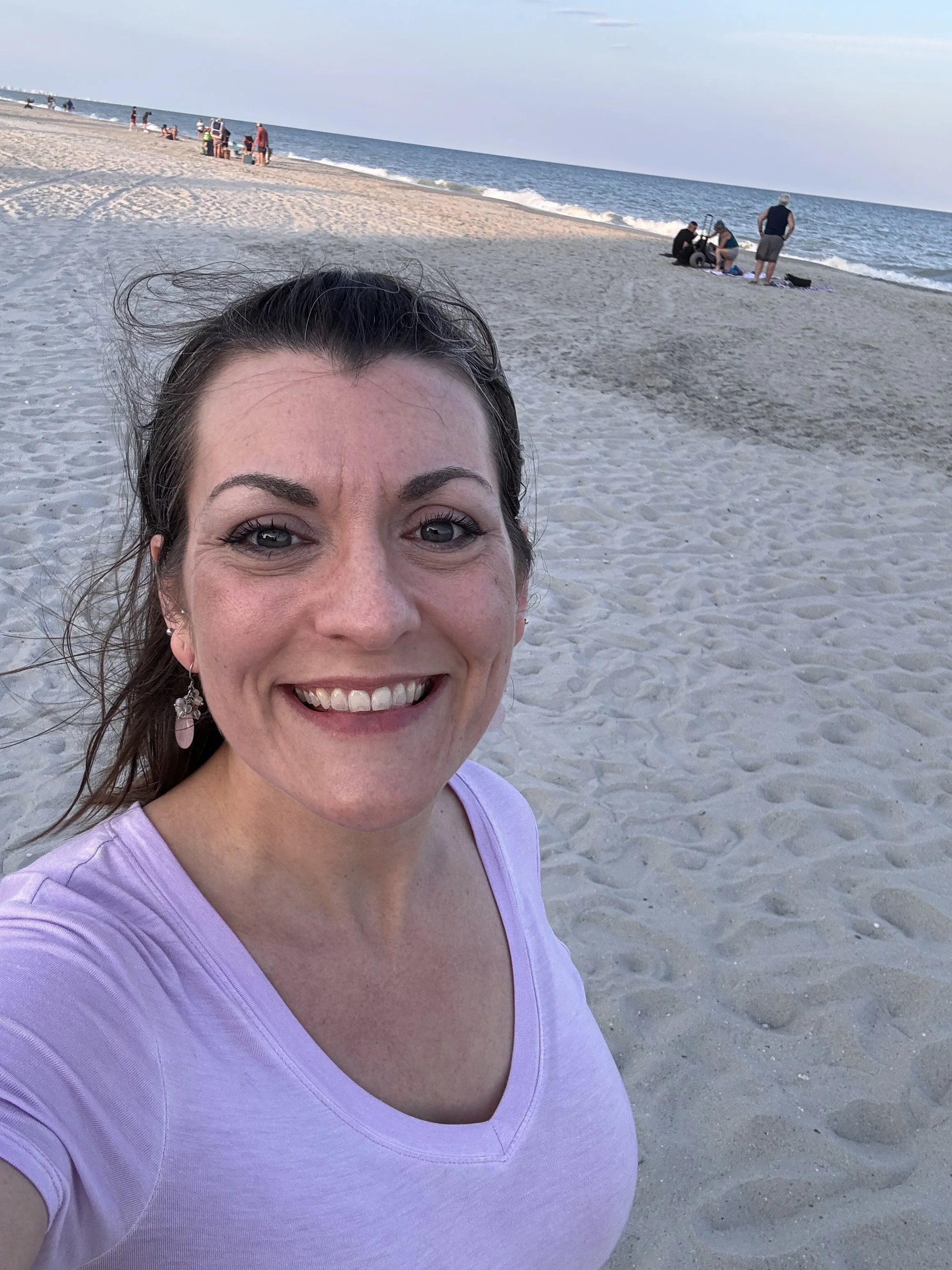 A woman smiling at the camera on a sandy beach with the ocean in the background.