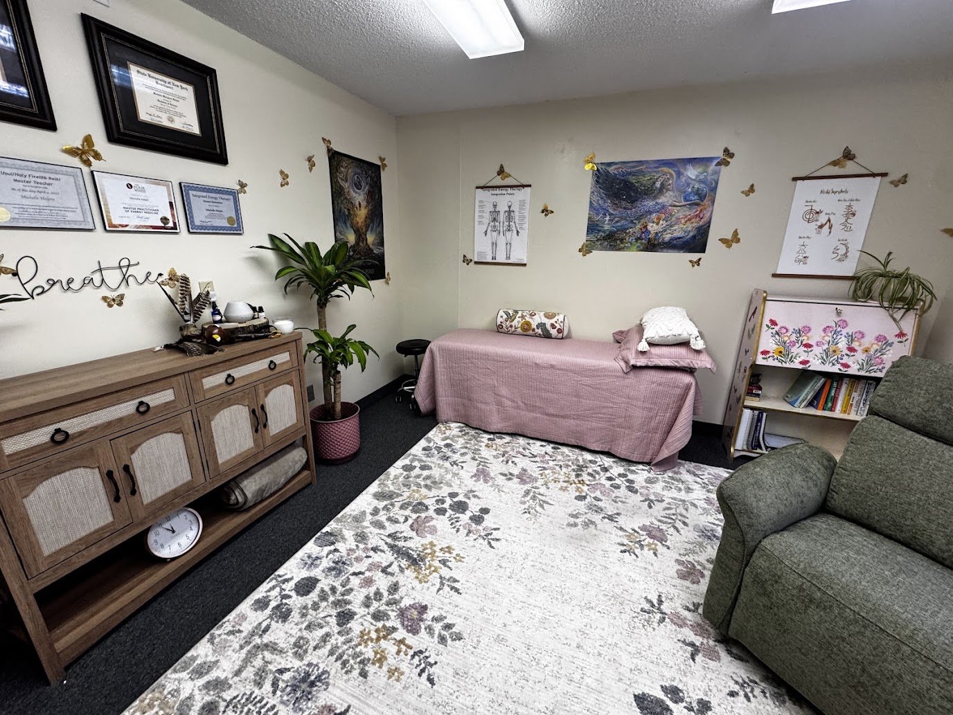 A therapy or counseling room with a green couch, a wooden cabinet, and a pink-covered massage table. Decor includes framed certificates, paintings, butterfly wall decals, a plant, and a bookshelf with books.