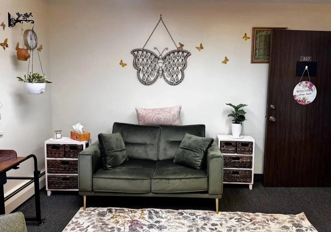 Living room style office with a dark green sofa, two side tables with baskets, potted plant, hanging butterfly wall decor, and welcome sign on the door.