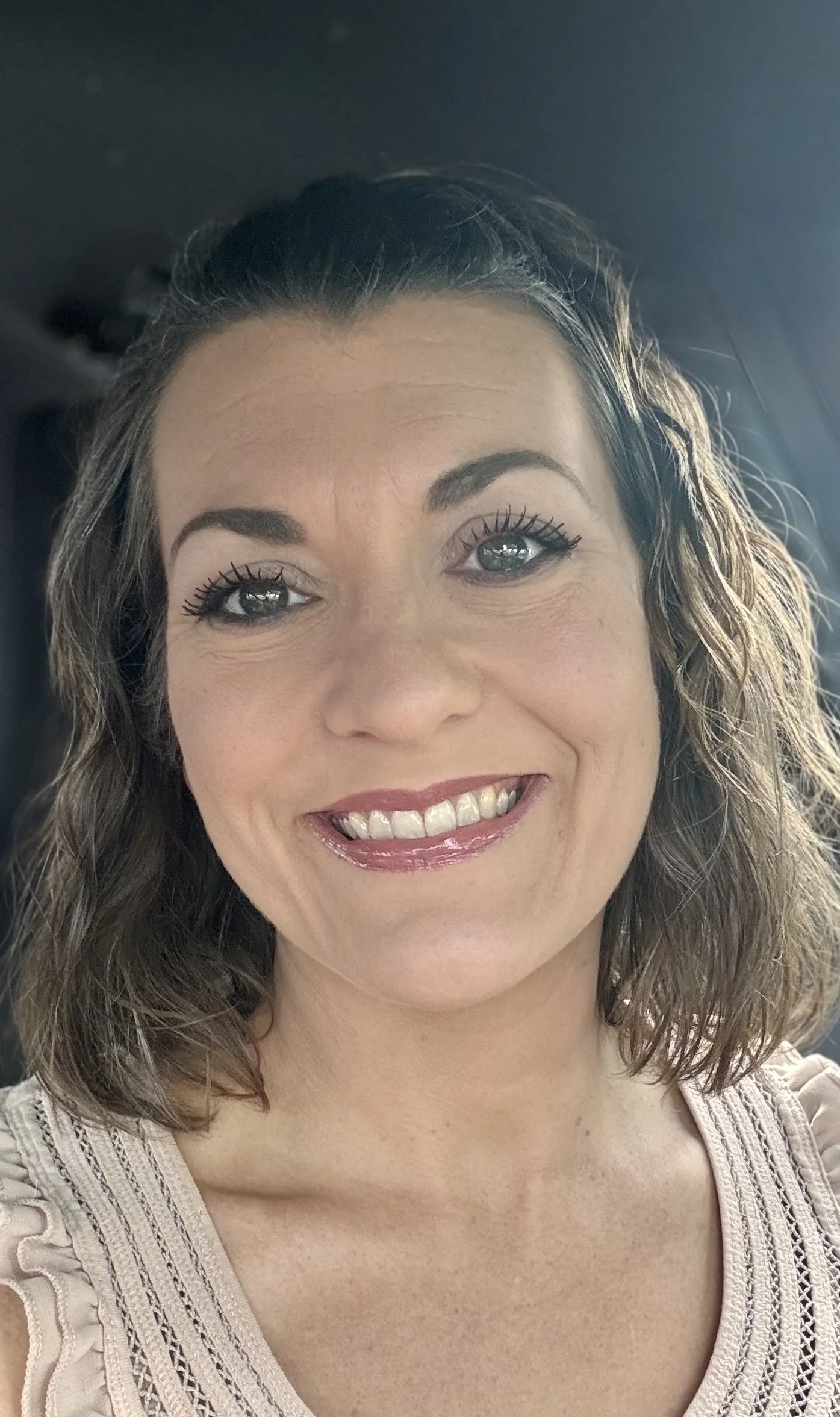 Smiling woman with curly hair and hazel eyes, wearing a beige top with lace details, indoors with a blurred background.