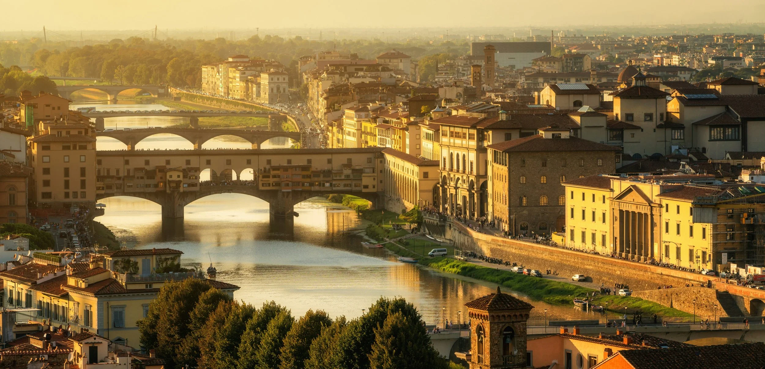 A cityscape of Florence, Italy, during sunset with historic buildings, bridges over the Arno River, and lush green trees in the foreground. Experience Tuscany's charm