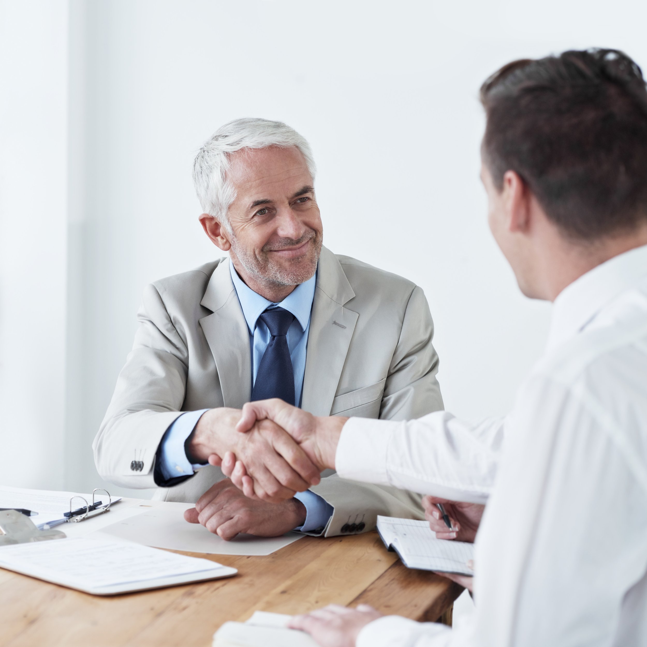 Two men in business attire shaking hands across a desk in an office setting.