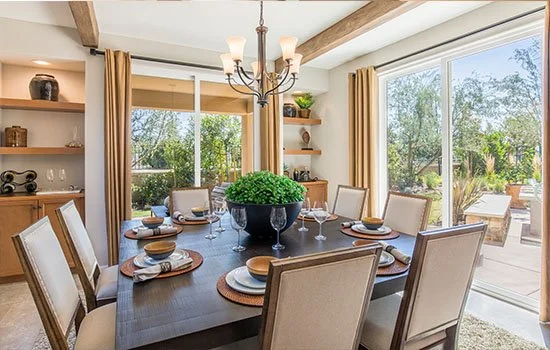 A dining room with a large wooden table set for six, featuring plates, bowls, and glasses, with a black vase of green flowers in the center, surrounded by cream-colored chairs. Large windows and sliding glass doors reveal a backyard with trees and outdoor furniture. A chandelier hangs above the table.